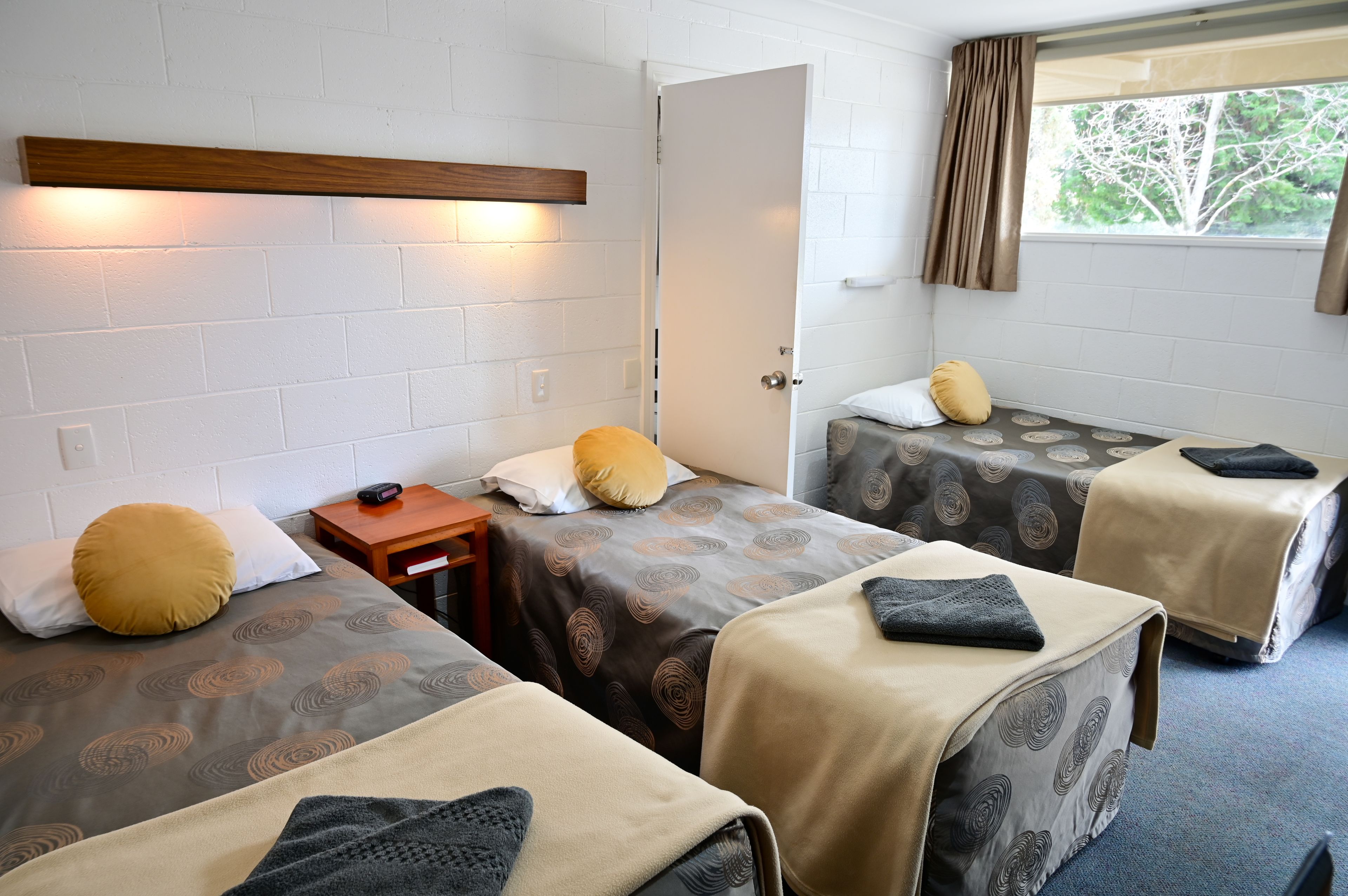 Une chambre avec trois lits simples, chacun recouvert d’une couvre-lit à motifs et agrémenté d’un oreiller. Au-dessus des lits, on trouve une tête de lit en bois, ainsi qu’une petite table en bois avec une chaise et une fenêtre avec des rideaux.