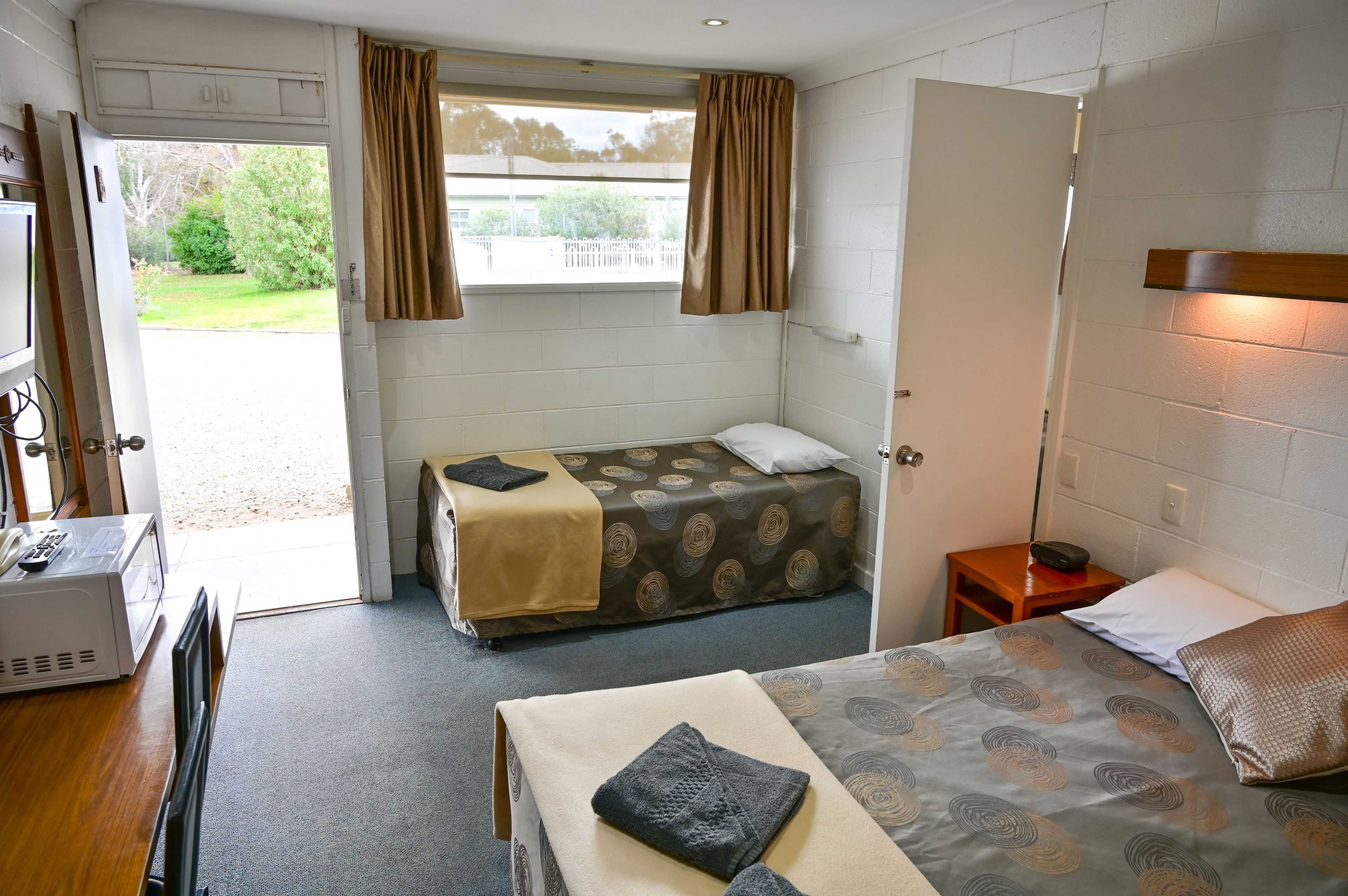 A single room with a bed, a desk, a chair, a window with curtains, and a door.