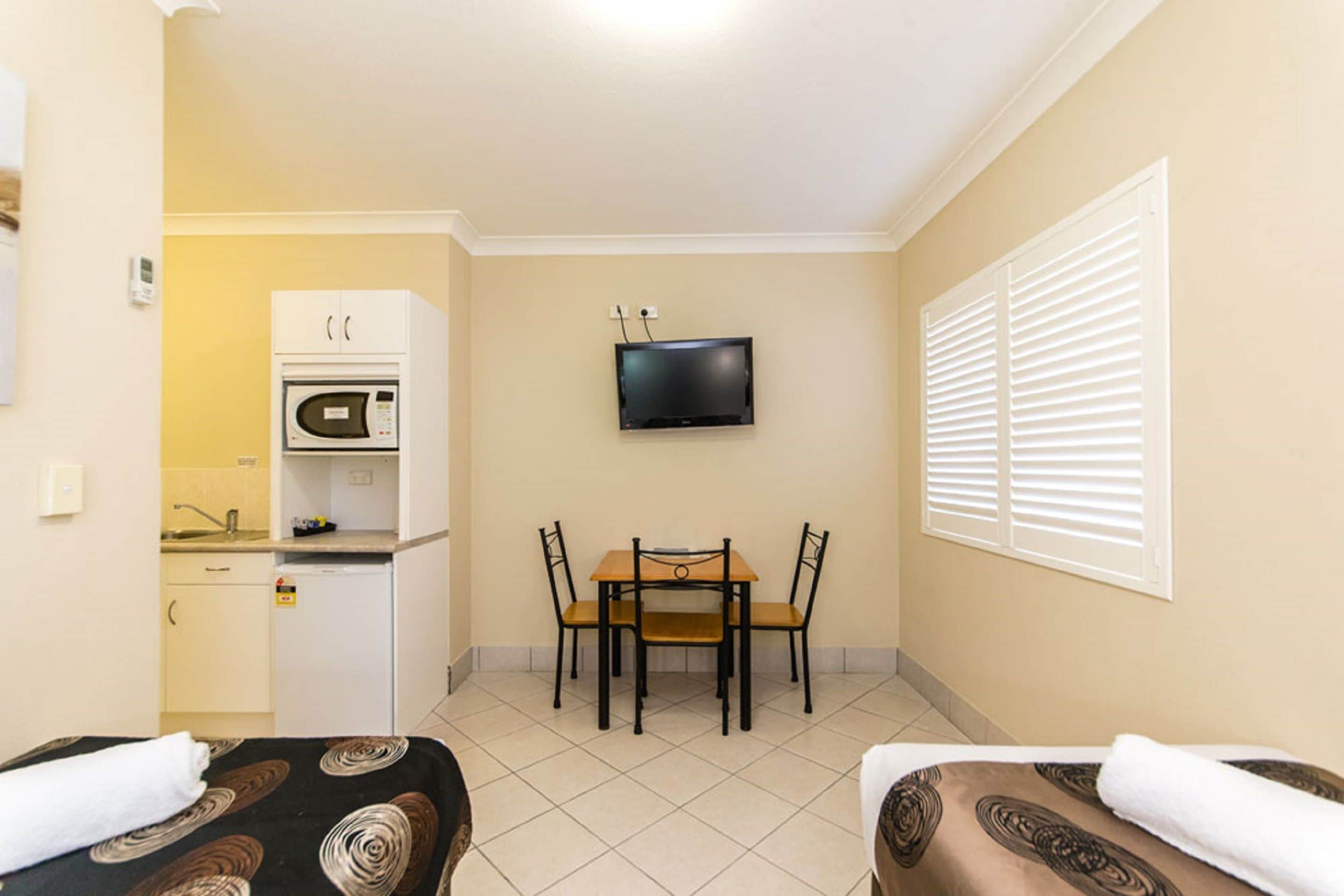 A hotel room with a bed, a small kitchenette, a dining table with chairs, and a TV mounted on the wall.