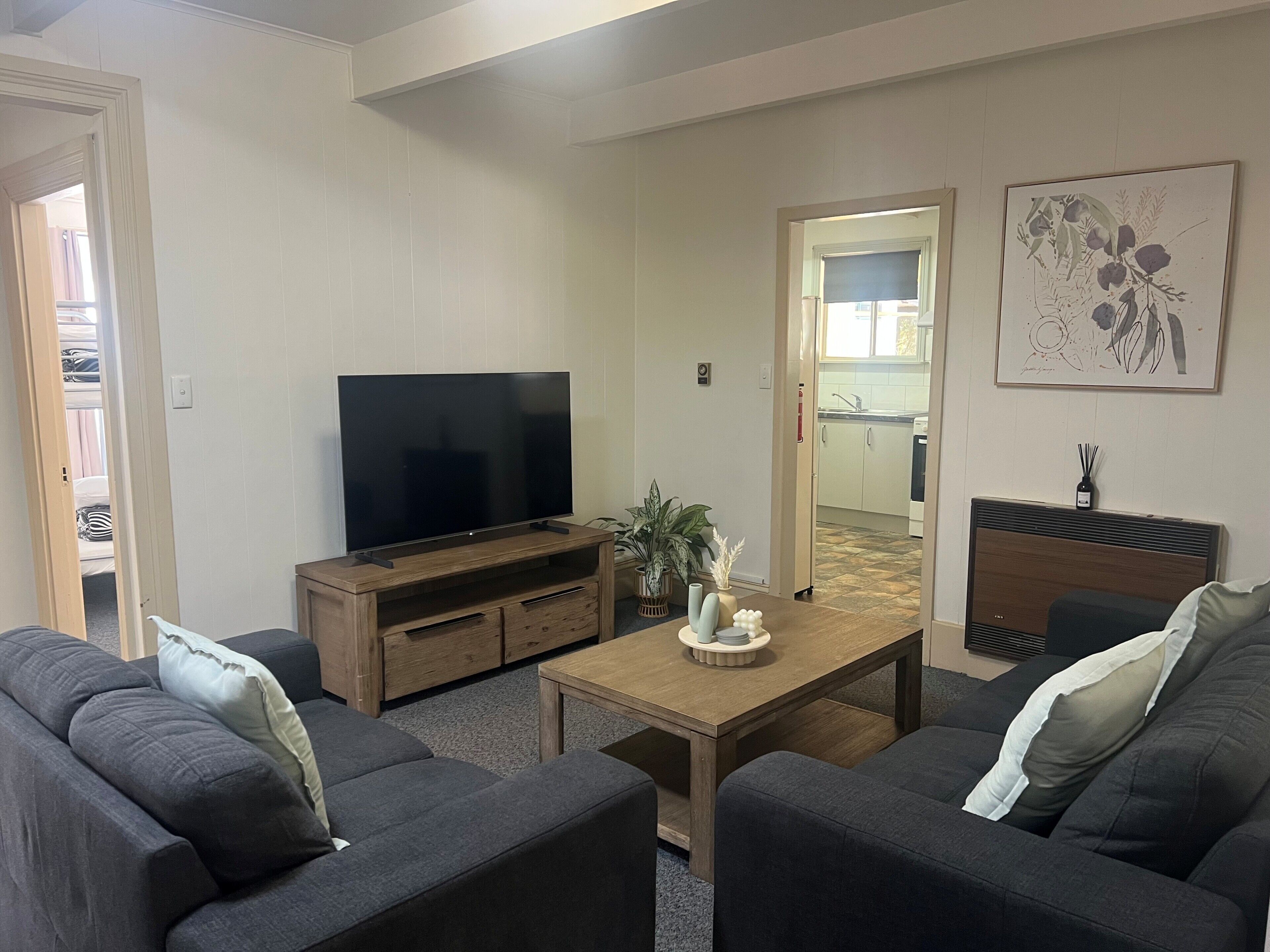 A living room with a flat-screen TV, wooden entertainment unit, dark sofas, a coffee table, and a kitchen in the background.