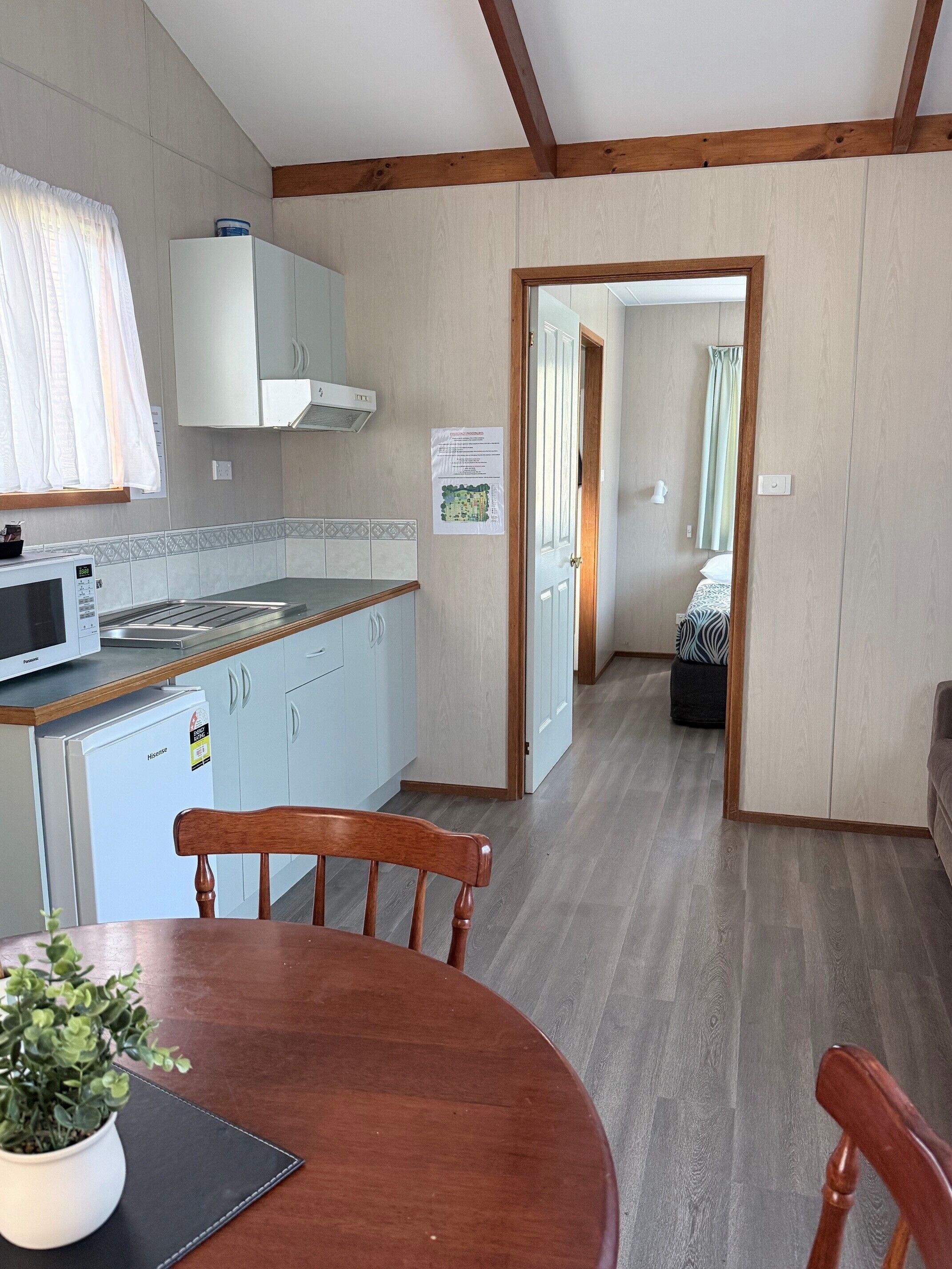 Una cucina moderna con forno a microonde, frigorifero e mobili in legno. C'è un tavolo da pranzo con sedie e una pianta in vaso. Un corridoio conduce a una camera da letto con un letto e una finestra.