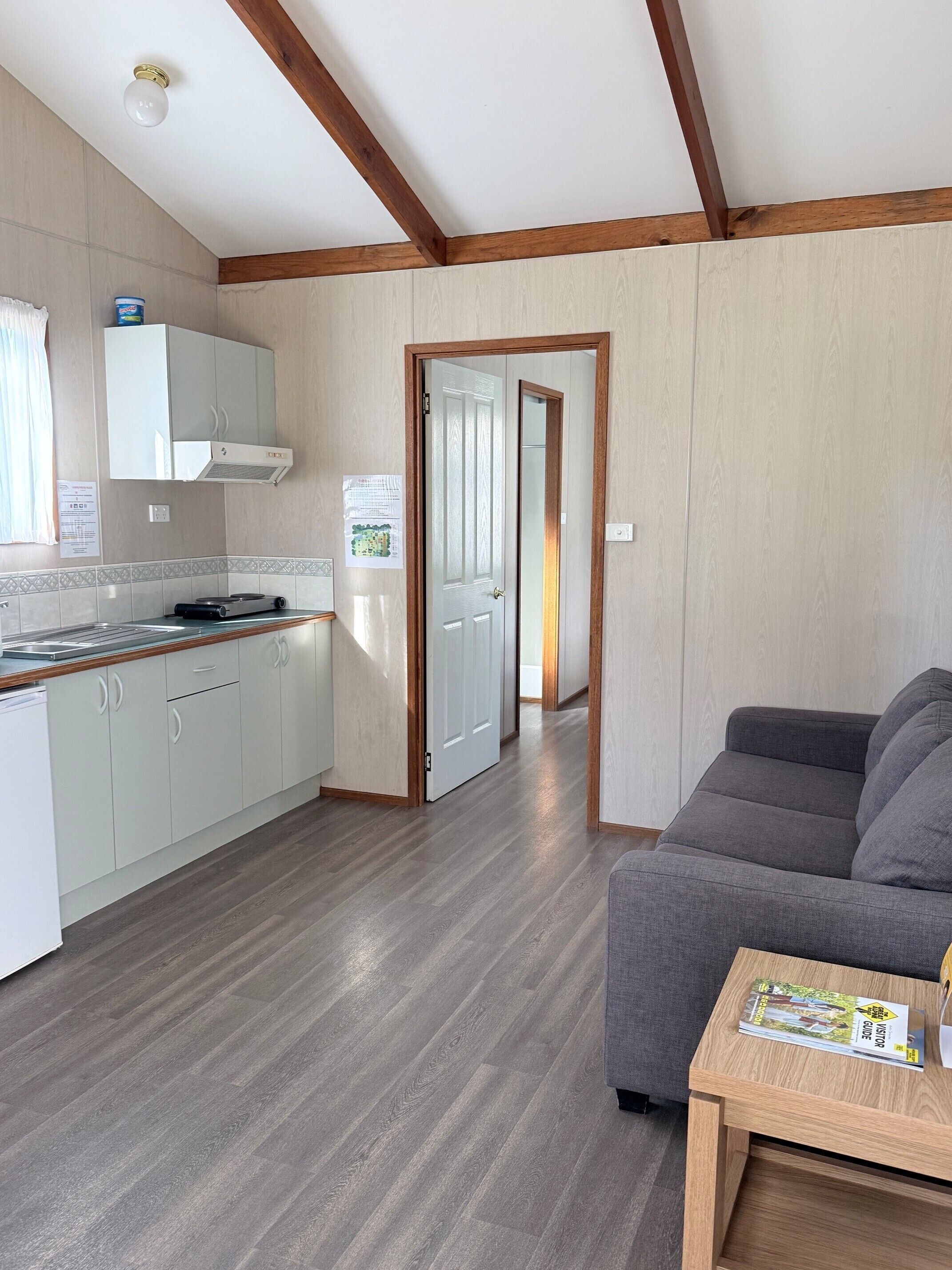A modern kitchen with white cabinets, a wooden countertop, and a built-in stove. There is a gray sofa and a wooden side table with a magazine. A hallway leads to a door.