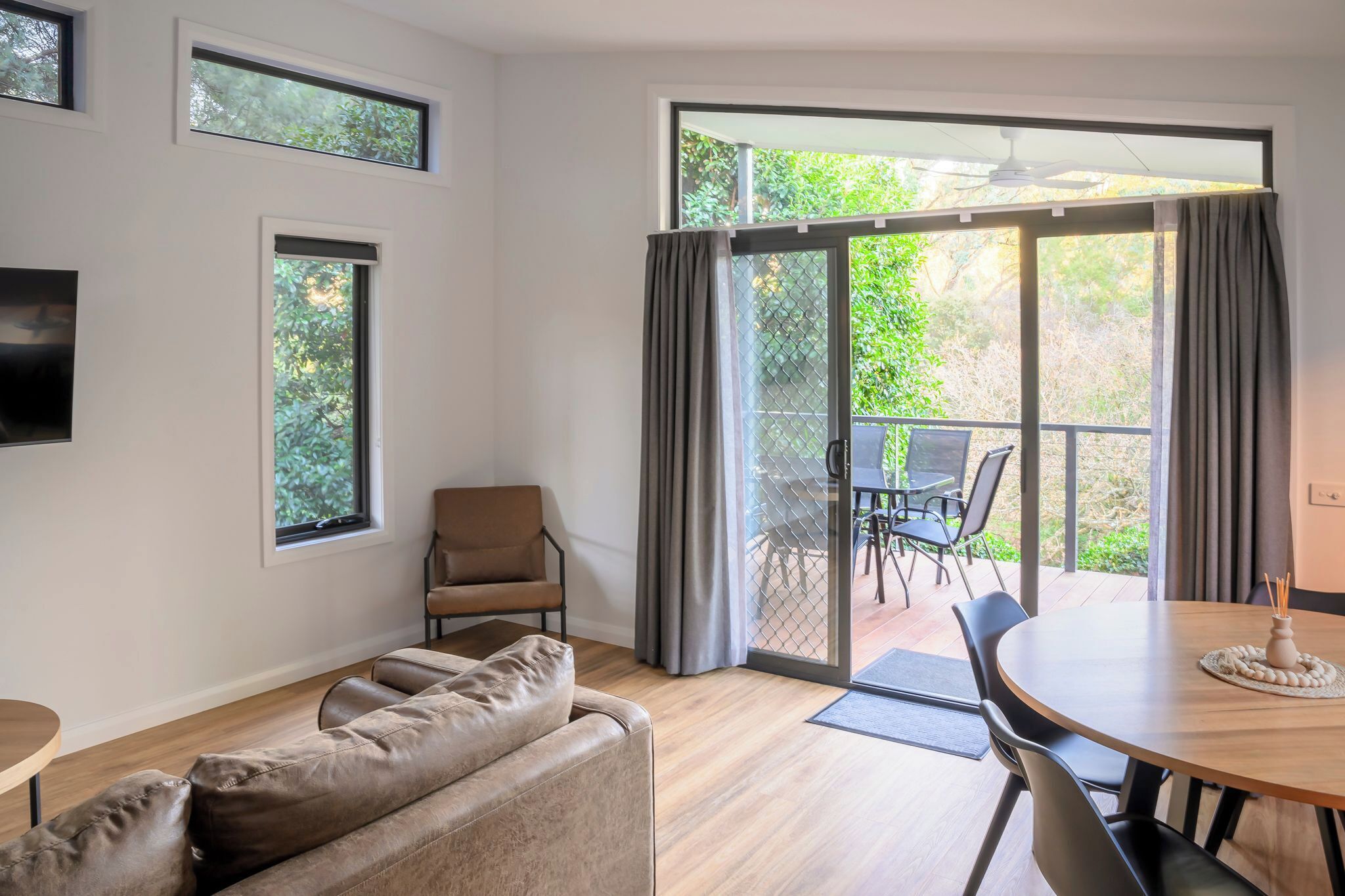 A modern living room with a sofa, a chair, a dining table, and a view of a balcony with outdoor furniture.
