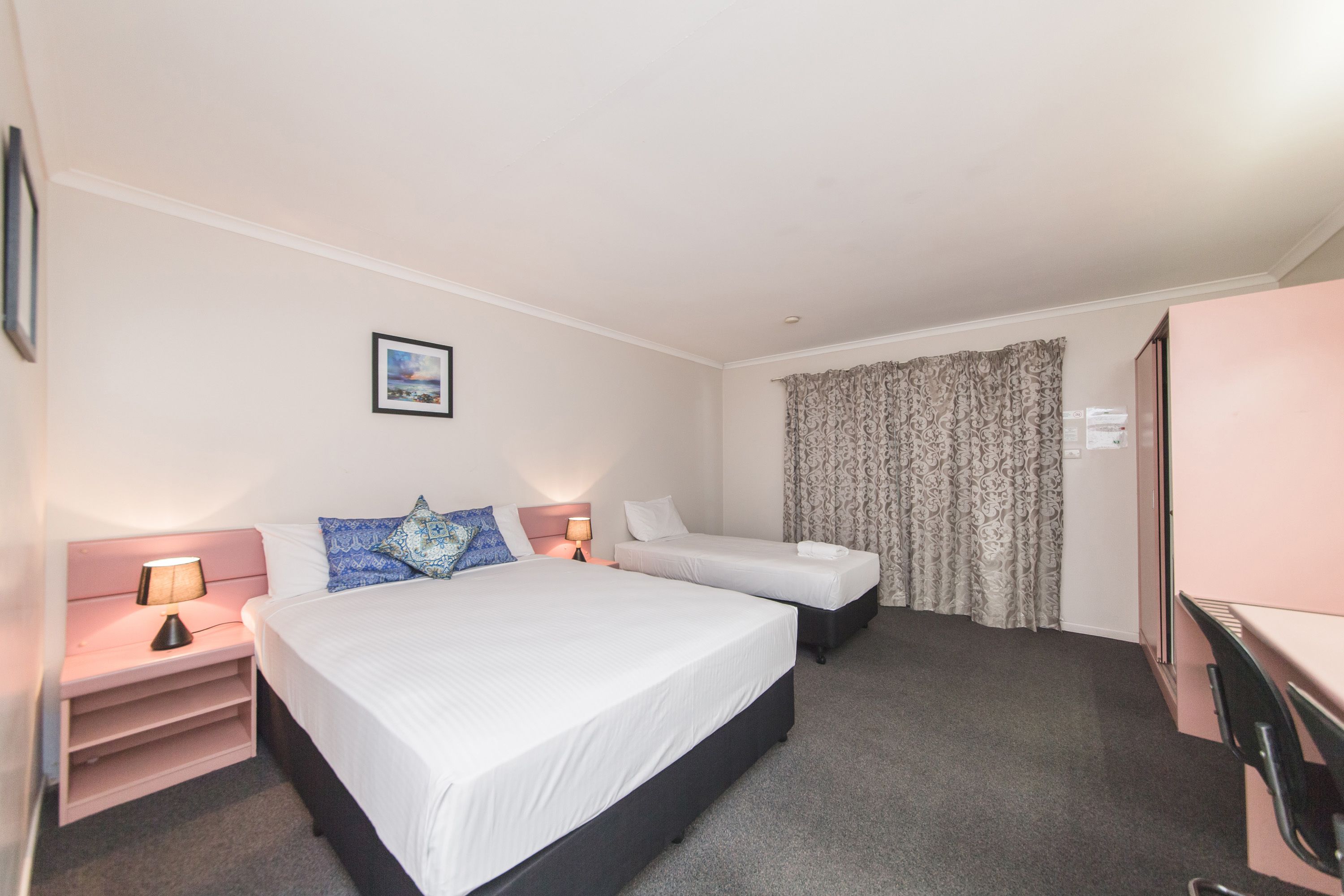A hotel room with a large bed, a smaller bed, a desk, and a wardrobe. There is a picture on the wall and a lamp on the nightstand.