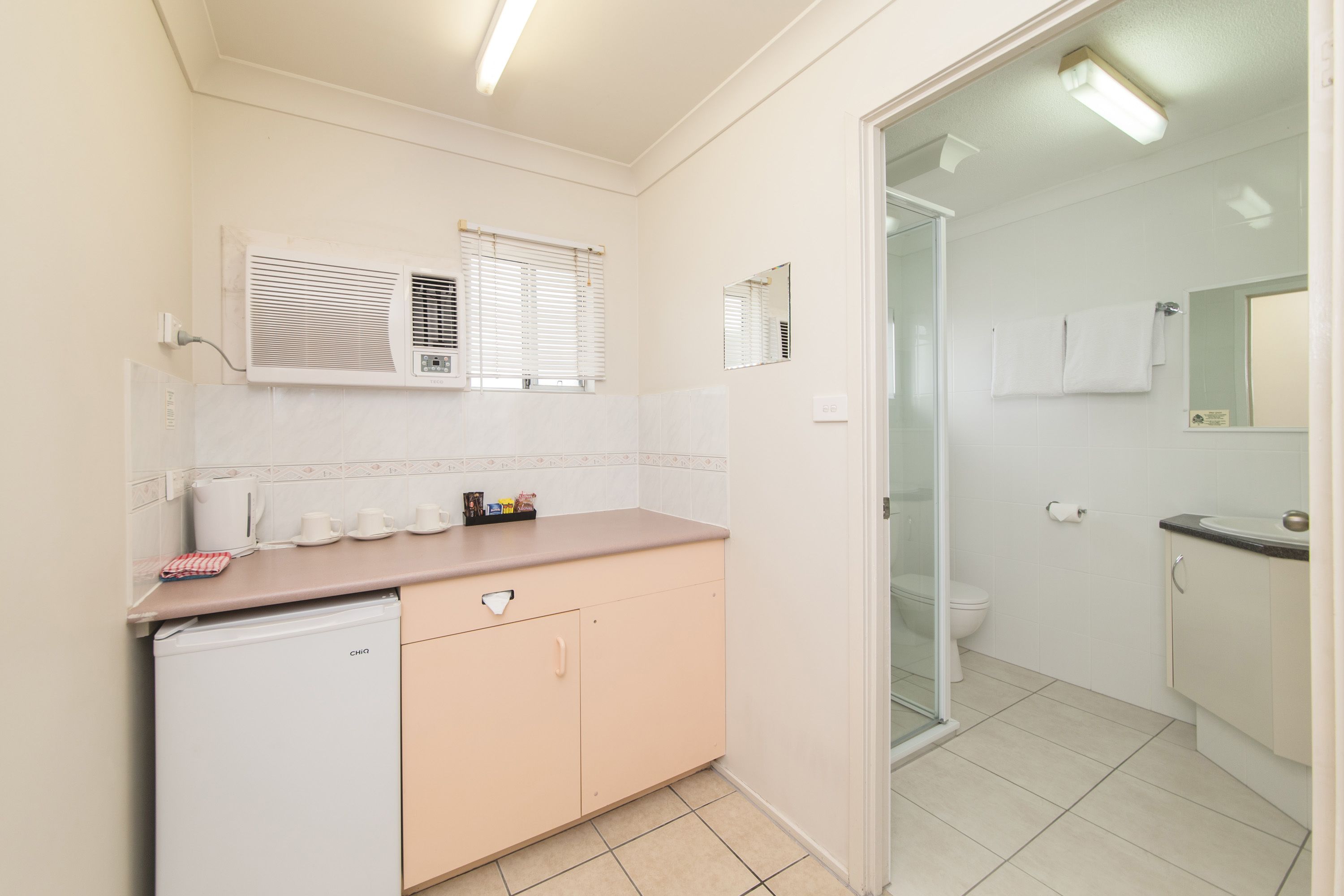 A compact bathroom with a toilet, sink, and a small refrigerator. There are two windows with blinds, and a mirror above the sink.