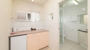 A compact bathroom with a toilet, sink, and a small refrigerator. There are two windows with blinds, and a mirror above the sink.