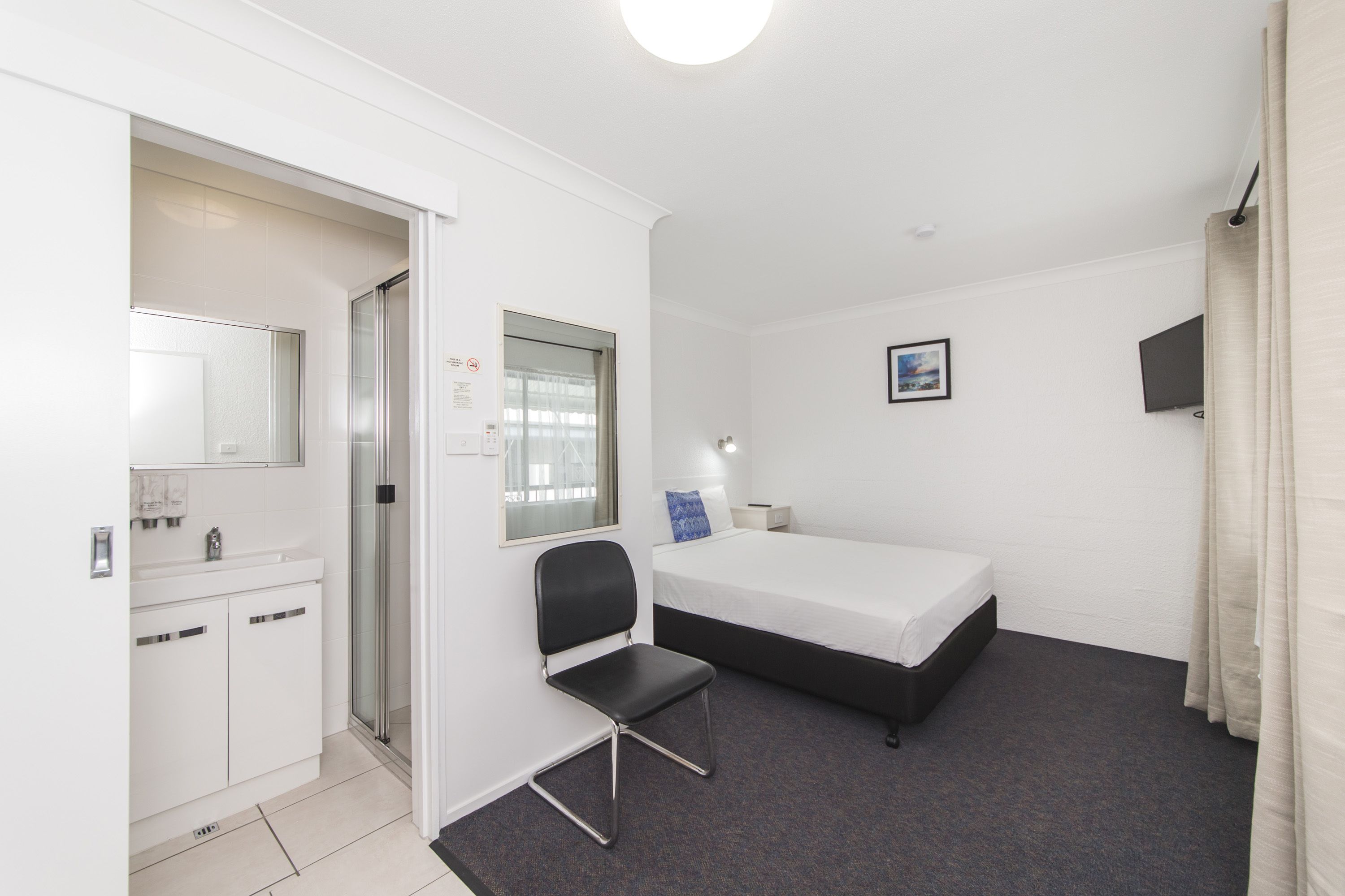 Une chambre d’hôtel compacte comprenant un lit simple, un petit bureau, une chaise, une fenêtre avec des rideaux, un téléviseur à écran plat et une salle de bain avec douche et lavabo.