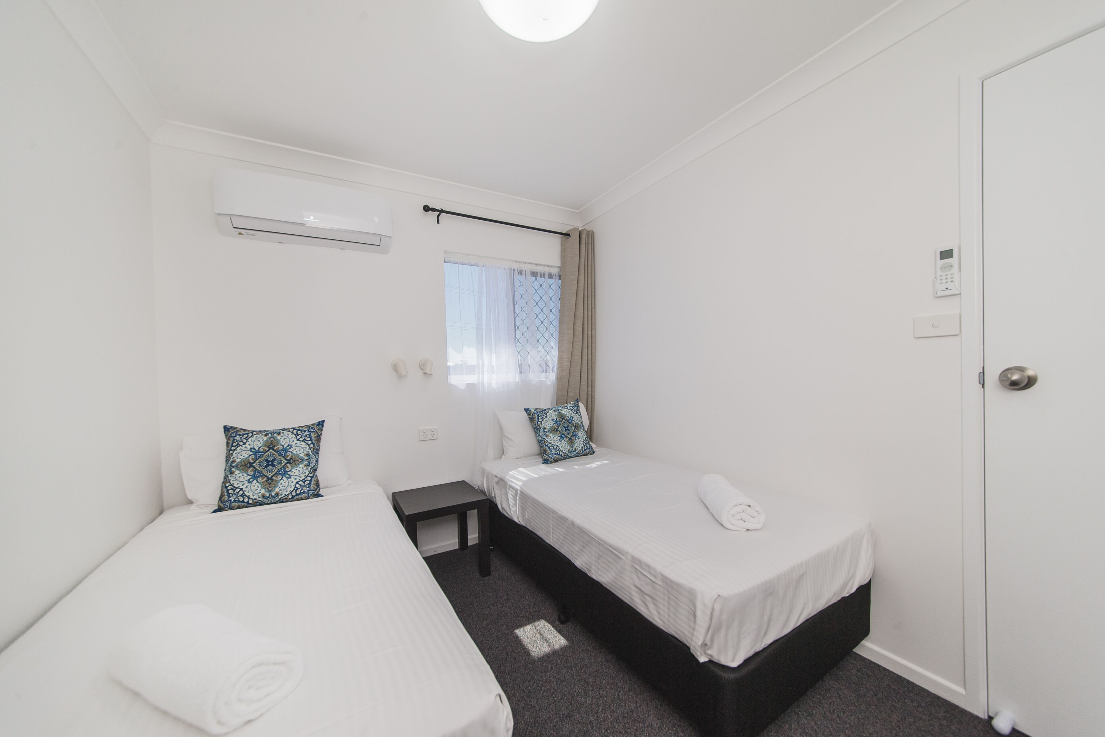 A small, clean room with two single beds, a small table, a window with curtains, and a door.