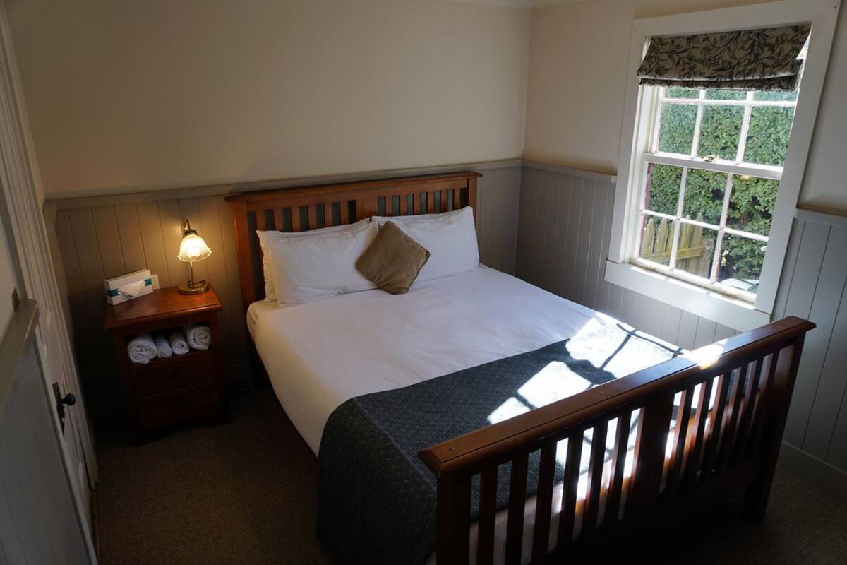 A bedroom with a bed, a nightstand with a lamp, a window with a view of greenery, and a wooden chair.