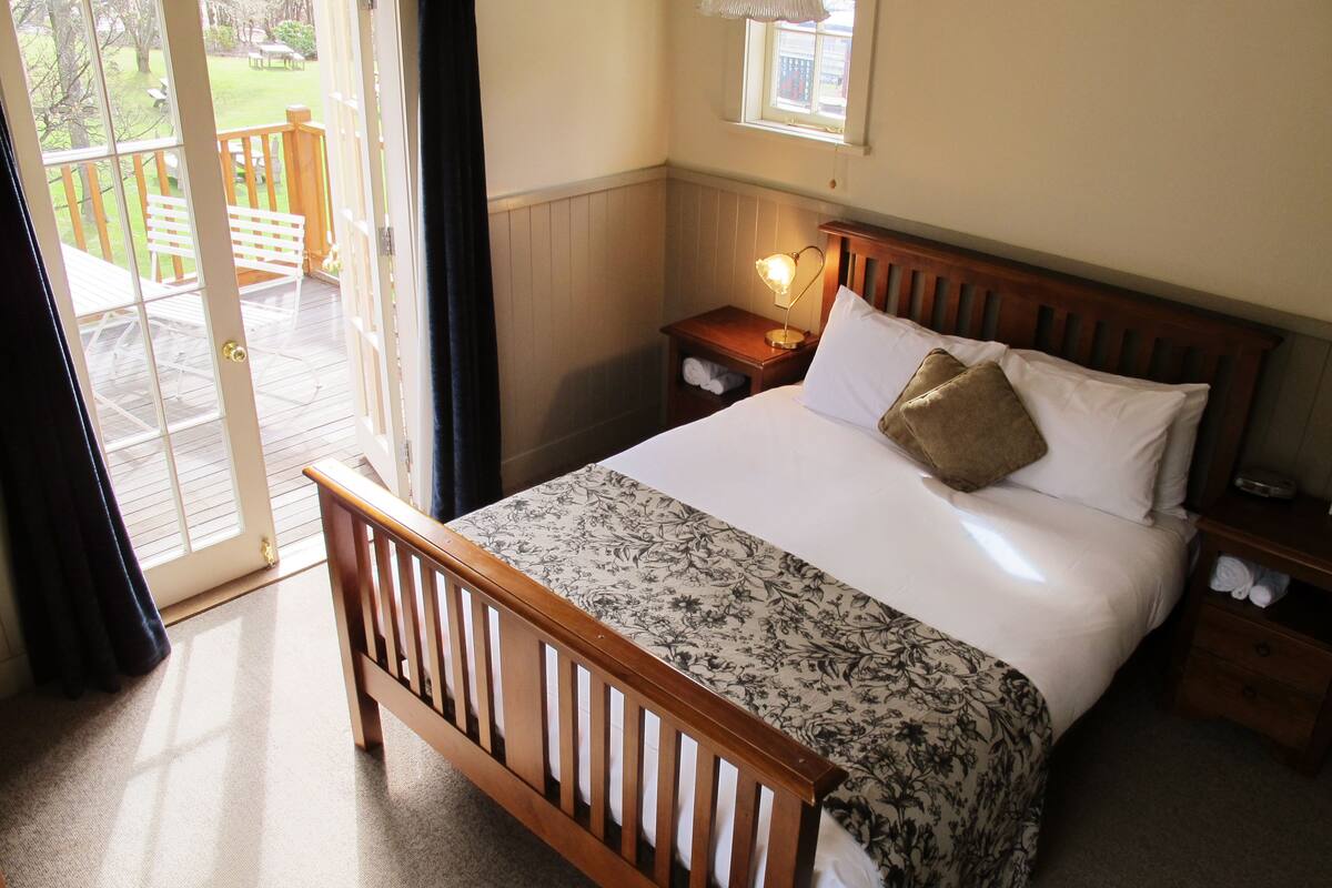 A bedroom with a wooden bed, a nightstand with a lamp, a window with a view of the outdoors, and a door leading to a balcony.