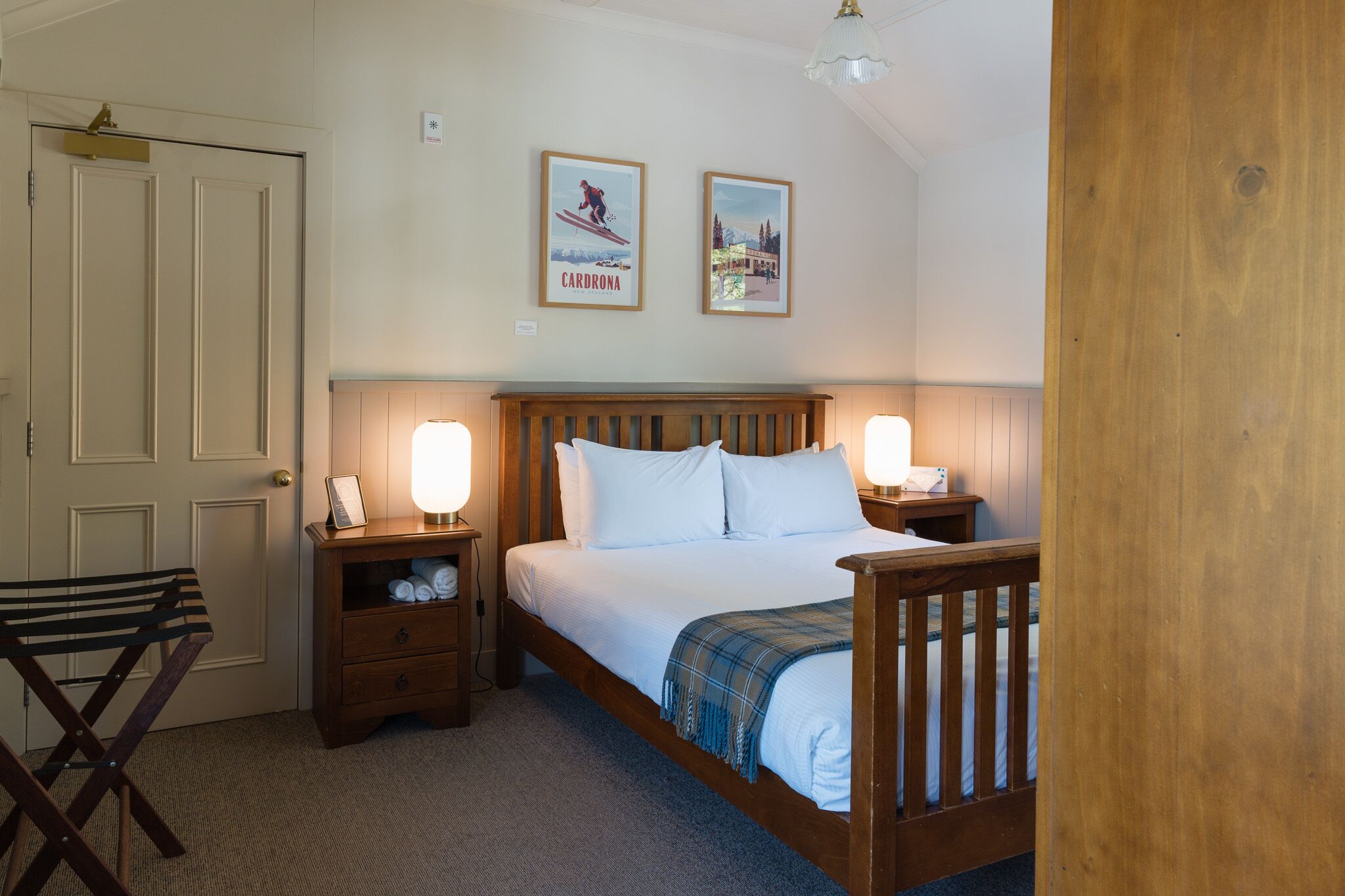 A neatly arranged bedroom with a wooden bed, bedside lamps, a small wooden nightstand, a chair, and two framed pictures on the wall.