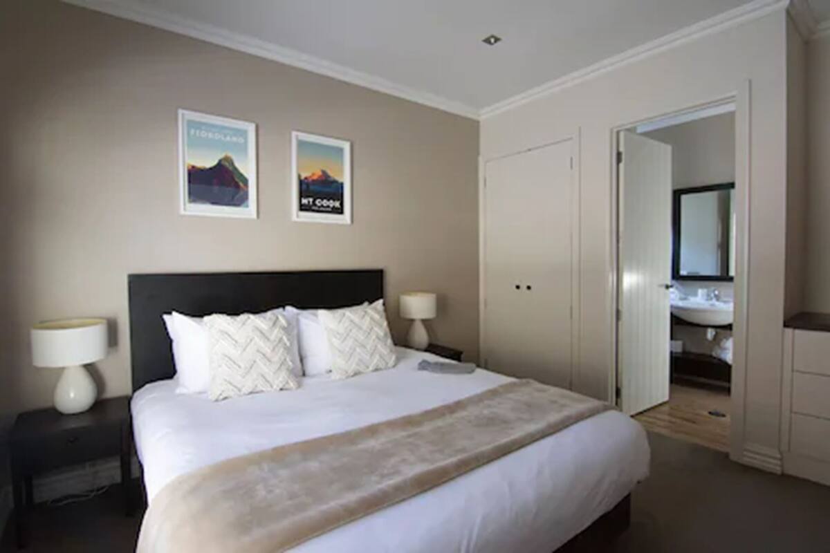 A bedroom with a bed, two nightstands, a headboard, and two wall-mounted lamps. There is a door leading to a bathroom.