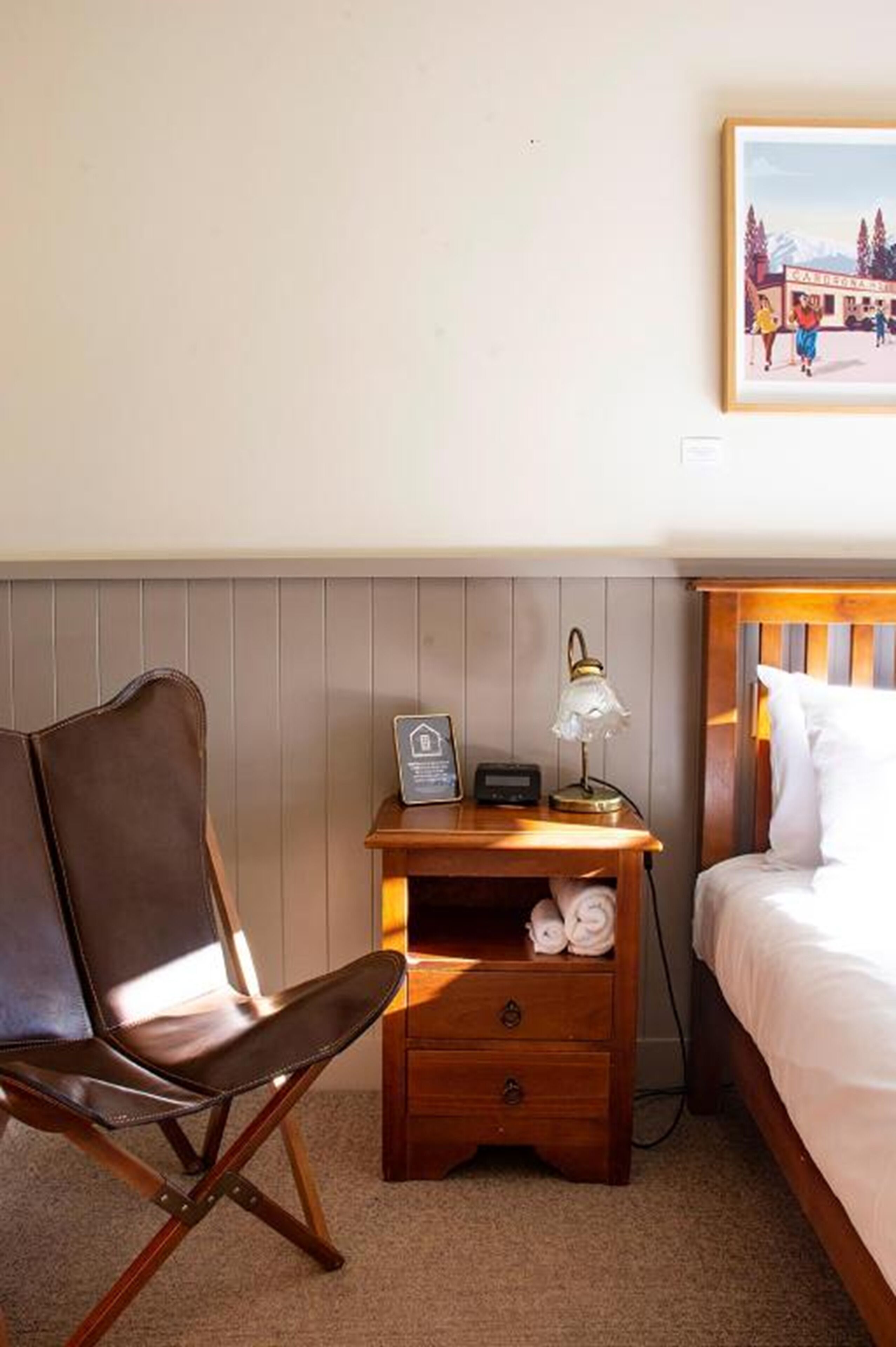 Habitación de hotel con una cama, una mesita de noche de madera con una lámpara, una silla y un cuadro colgado en la pared.