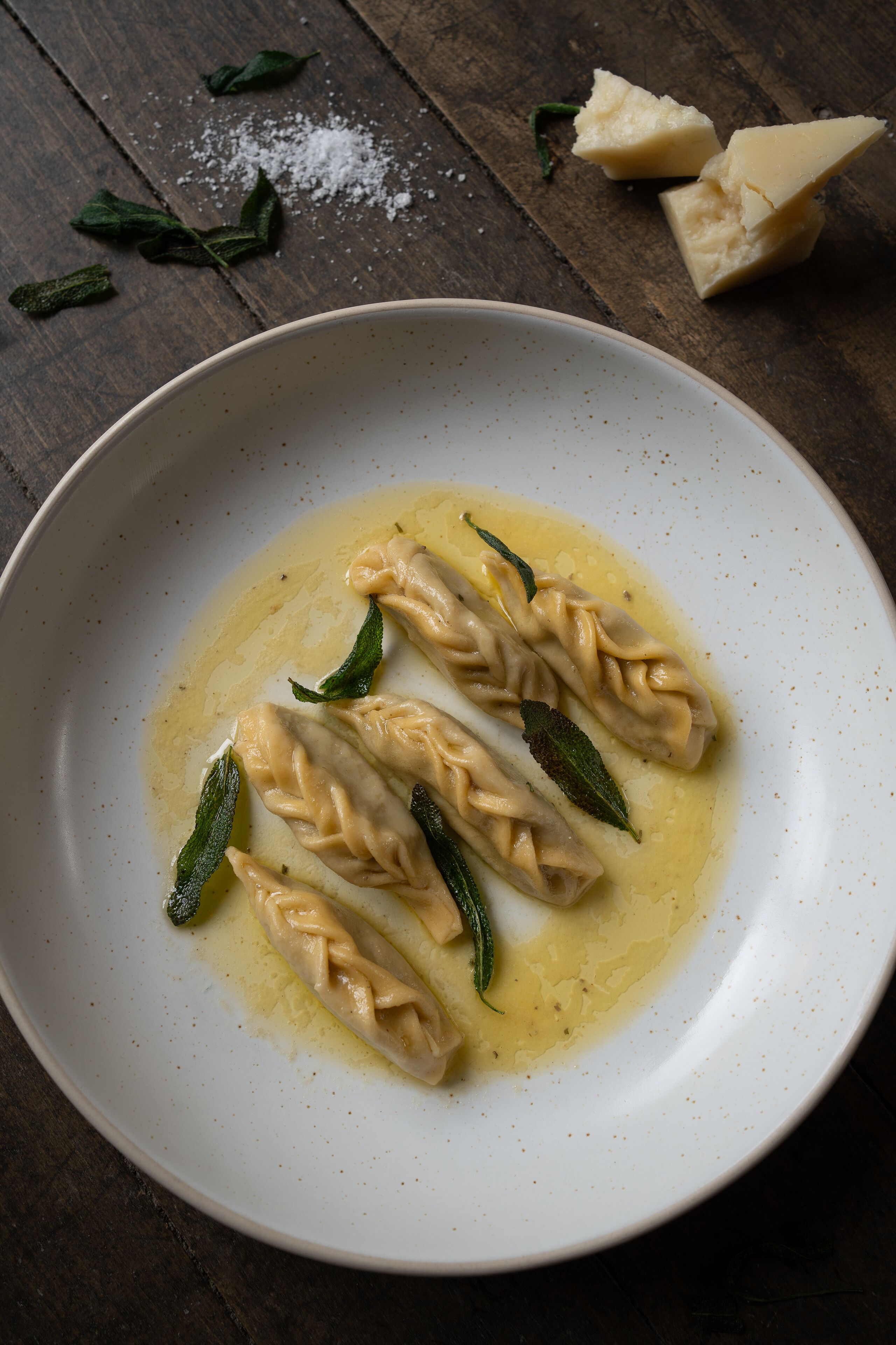A plate of ravioli with sage leaves, salt, and cheese on a wooden table.