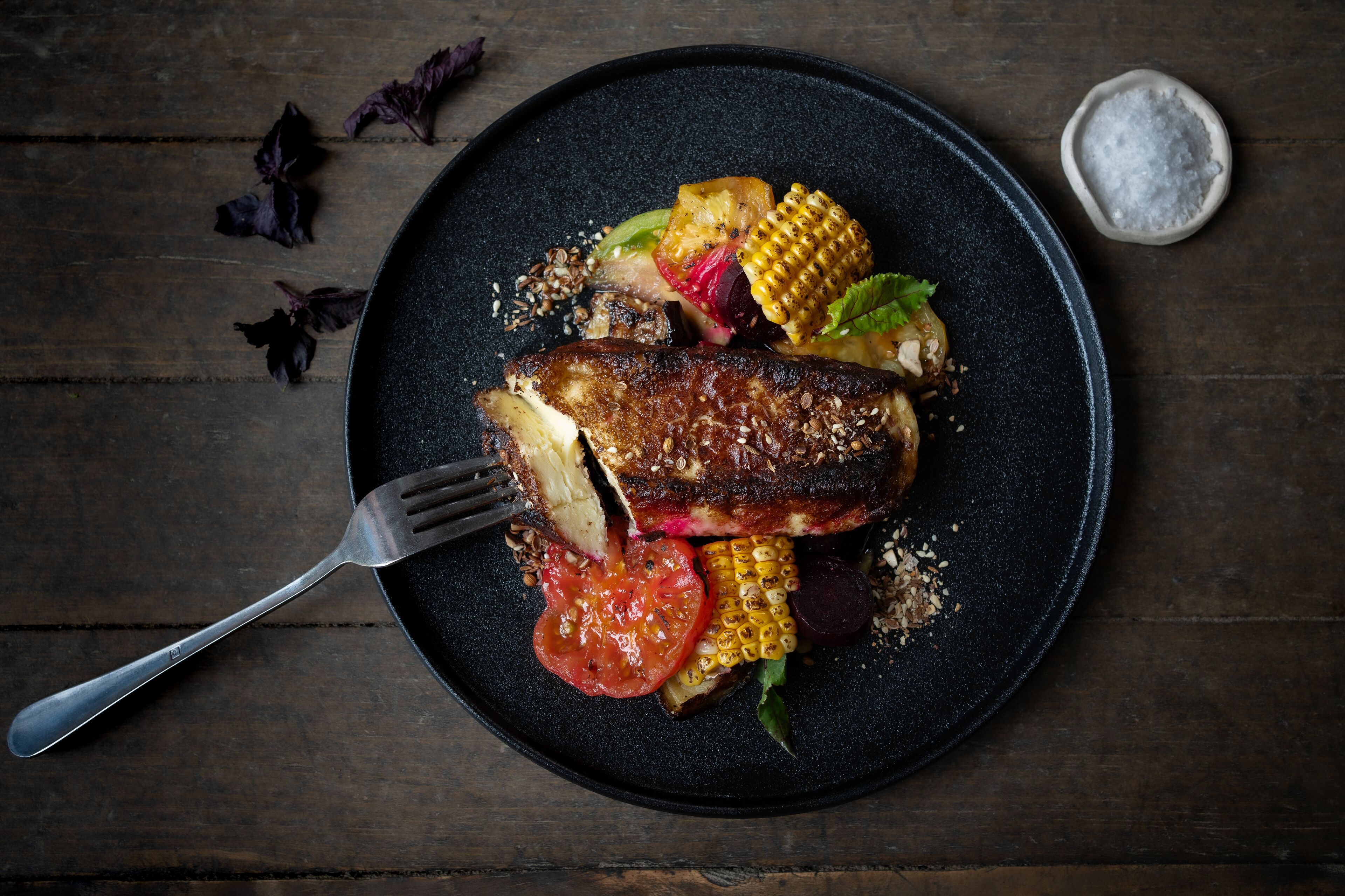 Une assiette contenant de la viande grillée, des légumes et une fourchette, le tout posé sur une assiette de couleur sombre.