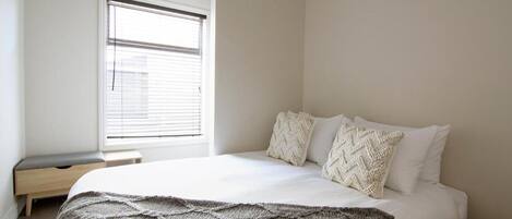 A bedroom with a bed, a window with blinds, a bedside table, and a wall-mounted shelf.