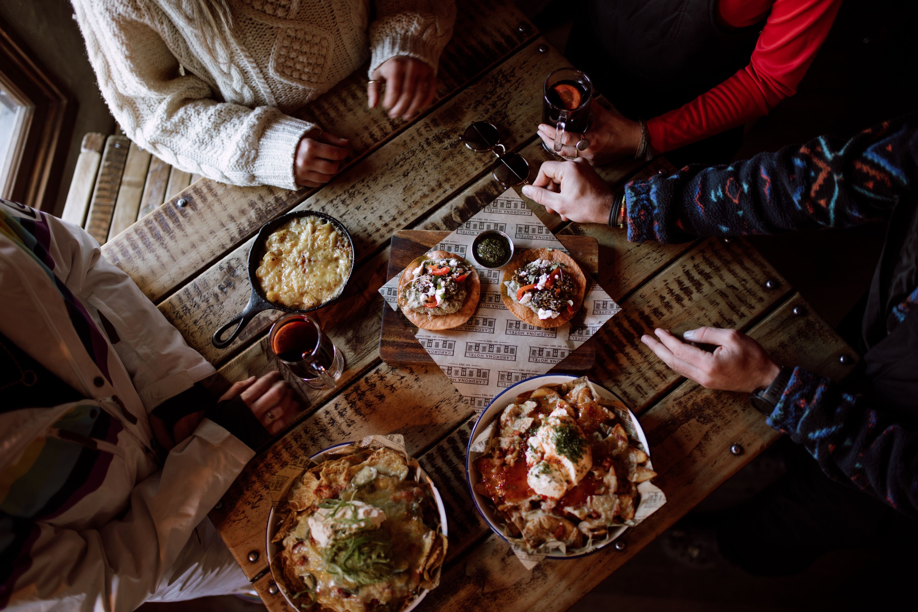Uma mesa de madeira com comida e bebidas, rodeada por pessoas.