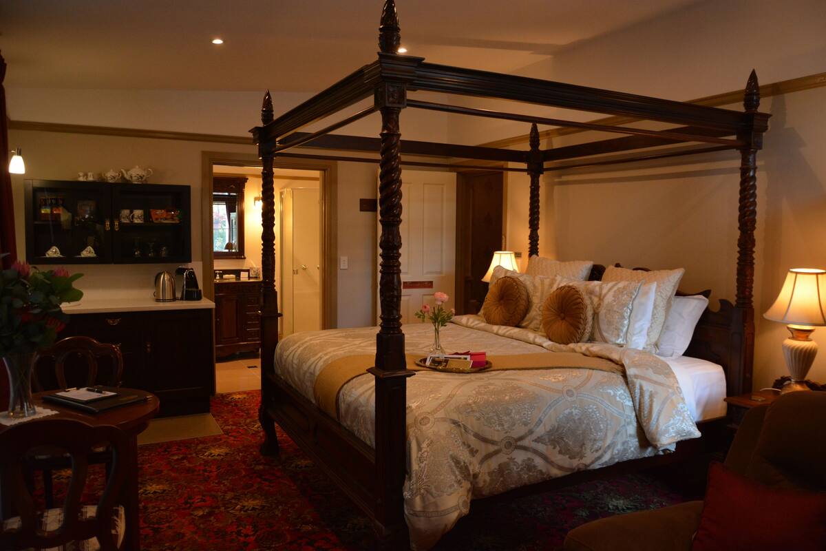 A four-poster bed with a floral bedspread, a bedside table with a lamp, and a small vase with flowers.