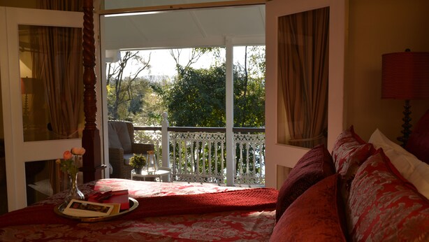 Ein Schlafzimmer mit einem großen Bett, einem Balkon mit weißer Brüstung und Blick auf Bäume und Himmel.