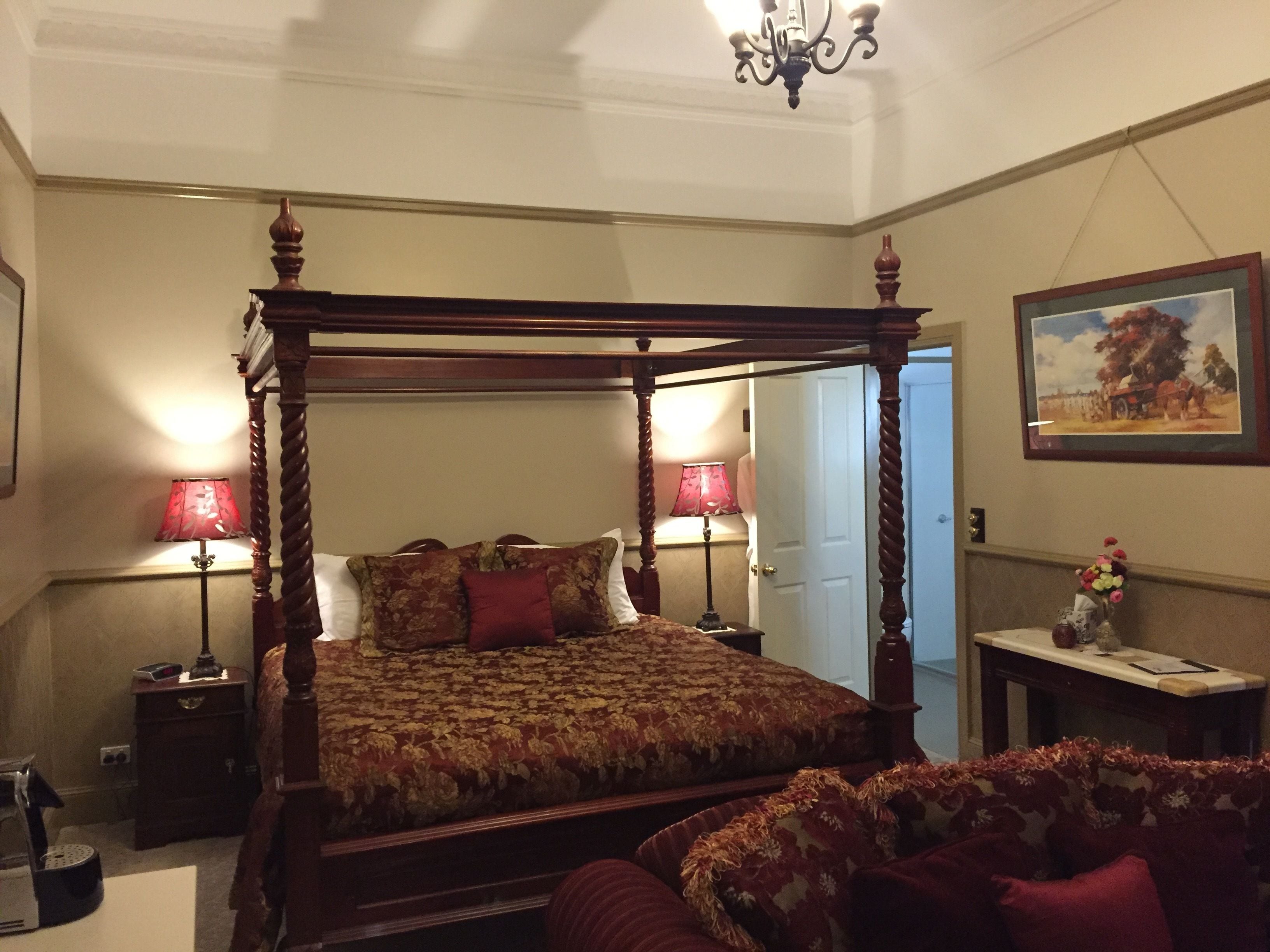 A four-poster bed with a floral bedspread and red pillows, two bedside lamps, a small table with a vase of flowers, and a framed painting on the wall.