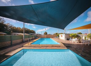 Una piscina rectangular con una zona sombreada arriba, rodeada por un patio de ladrillo y una cerca verde.