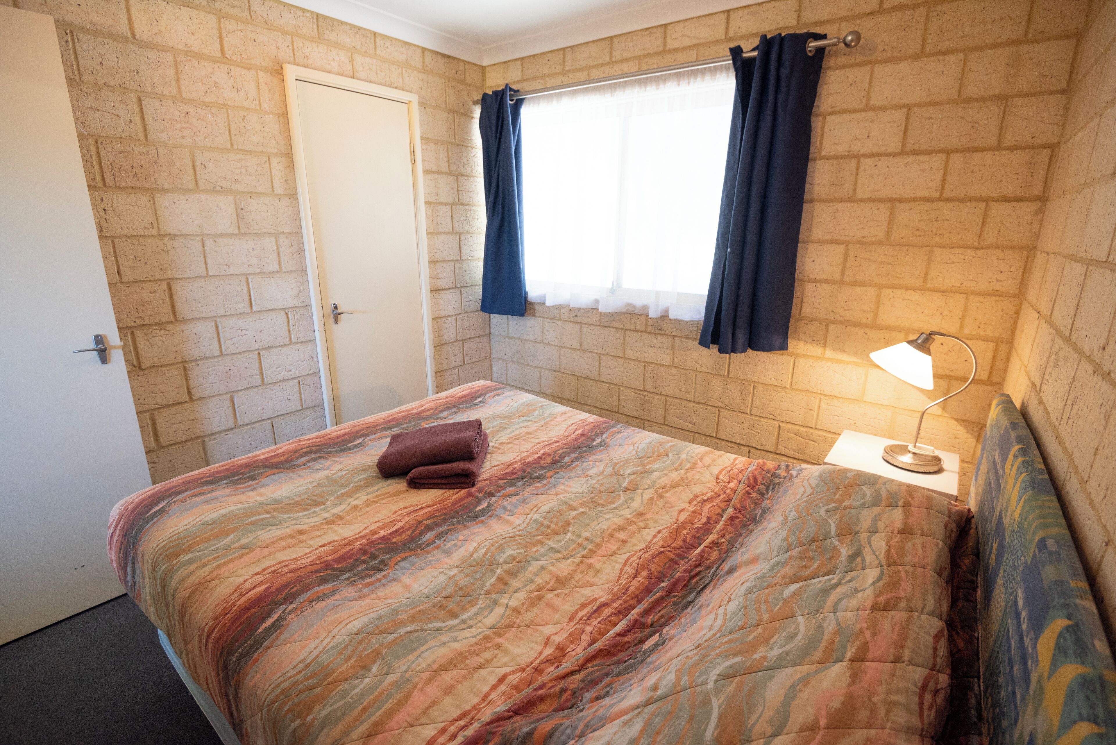 A bedroom with a brick wall, a window with curtains, a bed with a patterned bedspread, a bedside lamp, and a door.