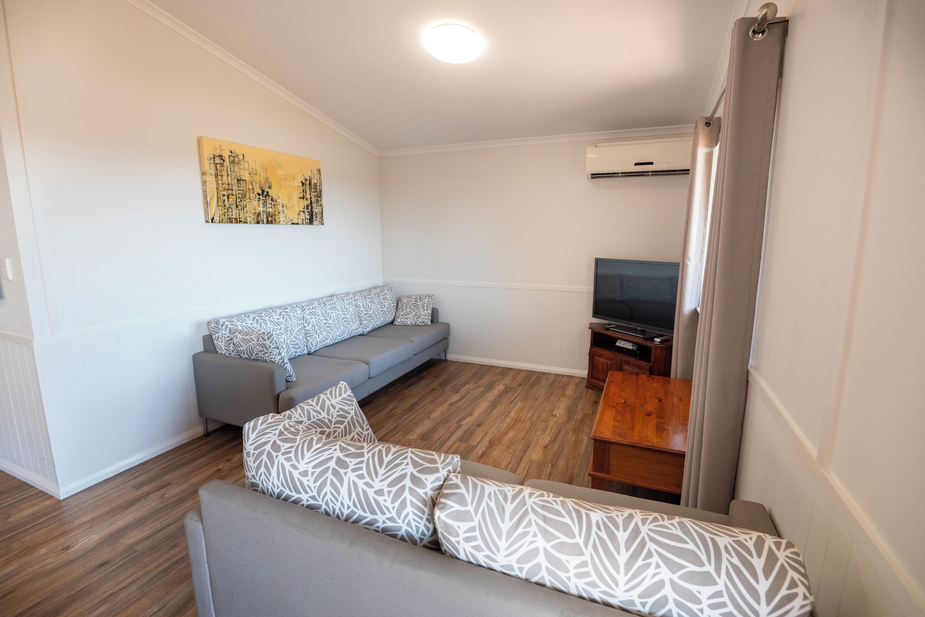 A modern living room with a grey sofa, patterned cushions, a wooden floor, a flat-screen TV, and a painting on the wall.