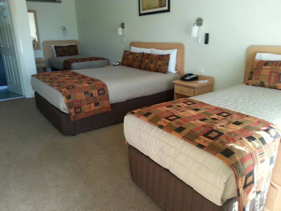 A hotel room with two beds, each with a patterned bedspread and pillows, a bedside table with a lamp, and a wall-mounted light fixture.
