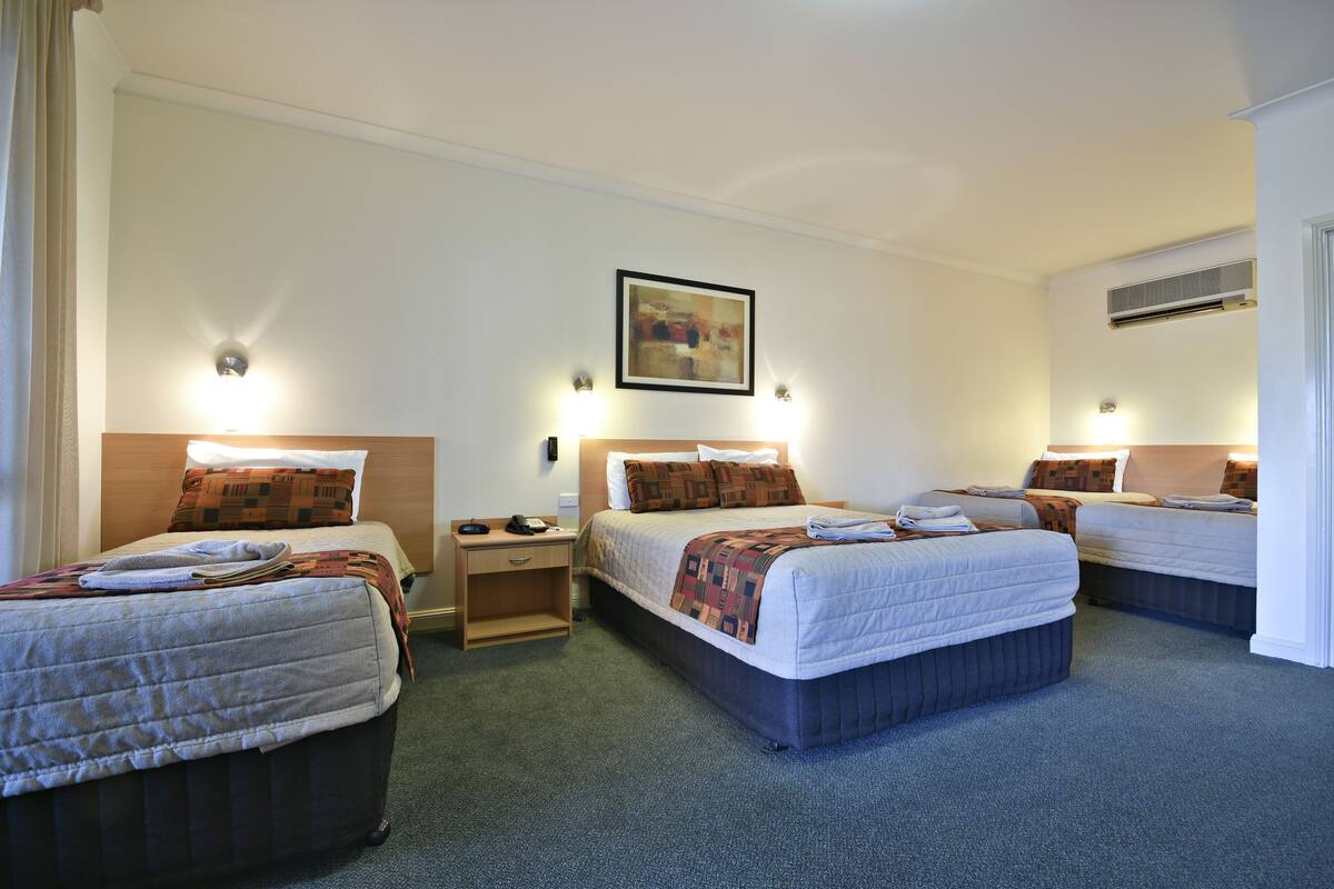 A hotel room with two single beds, a small desk, and a nightstand. There is a painting on the wall and an air conditioning unit above the beds.