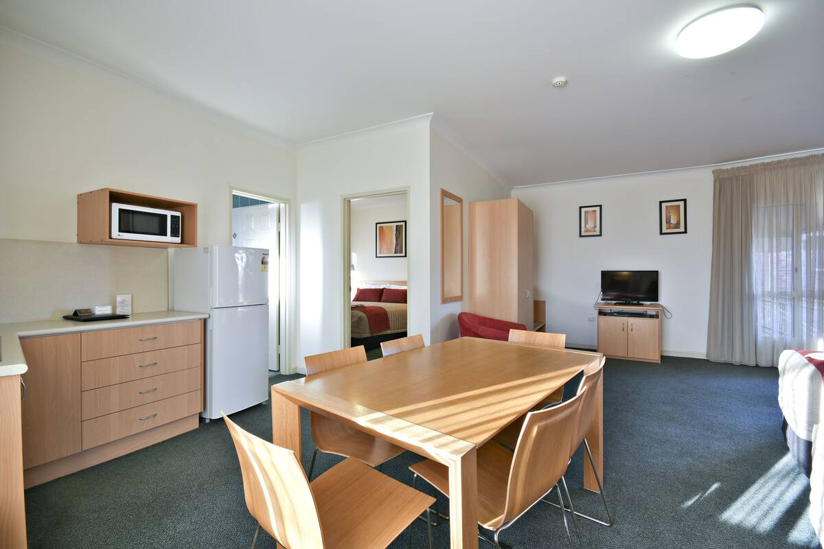 A compact hotel room with a kitchenette, a dining area, a TV, and a bed.