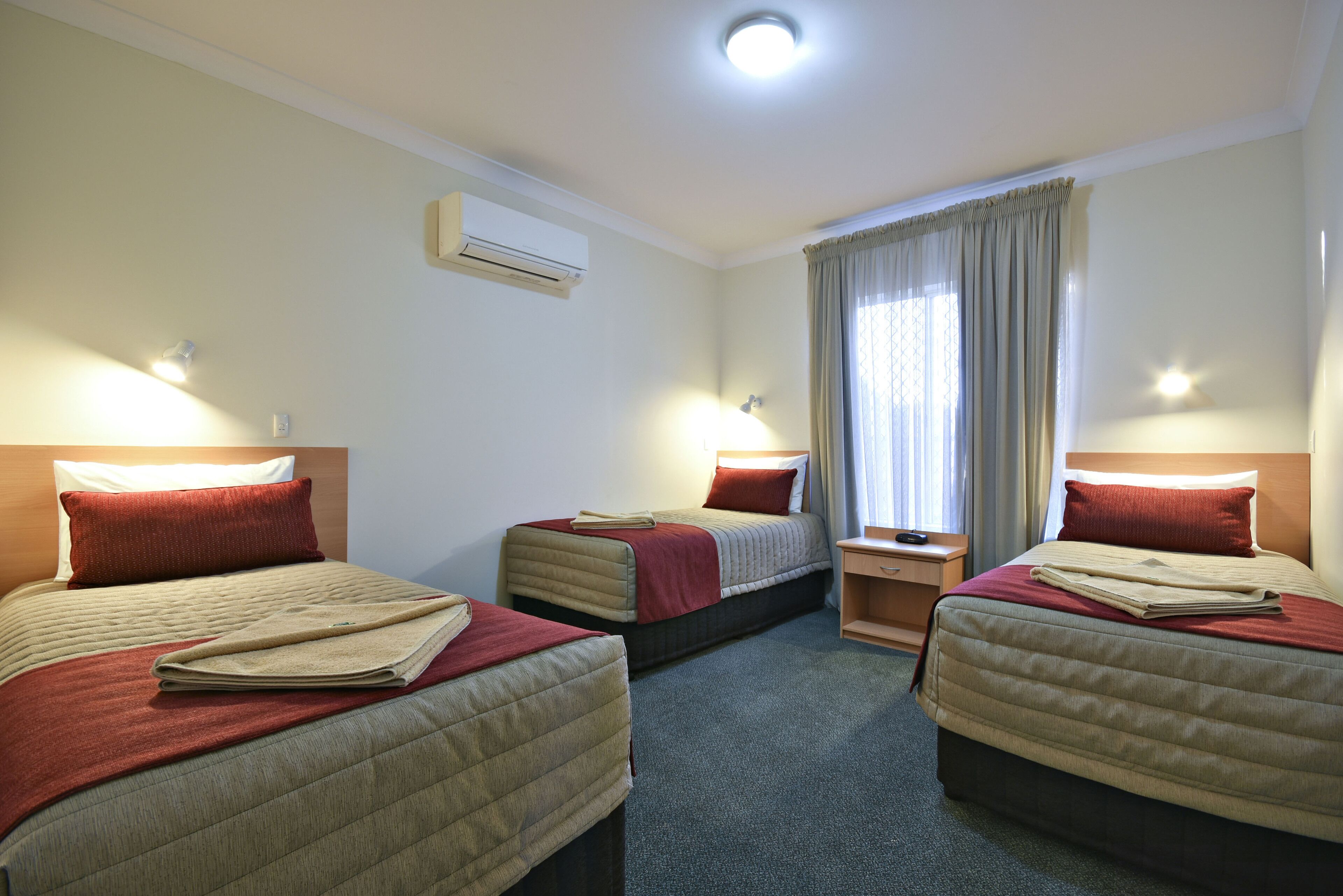 Habitación de hotel con tres camas, ventilador de techo, aire acondicionado y una ventana con cortinas.