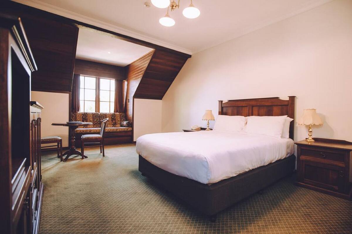 Een hotelkamer met een groot bed, twee nachtkastjes met lampen, een houten hoofdbord, een klein tafeltje met stoel, en een raam met gordijnen.
