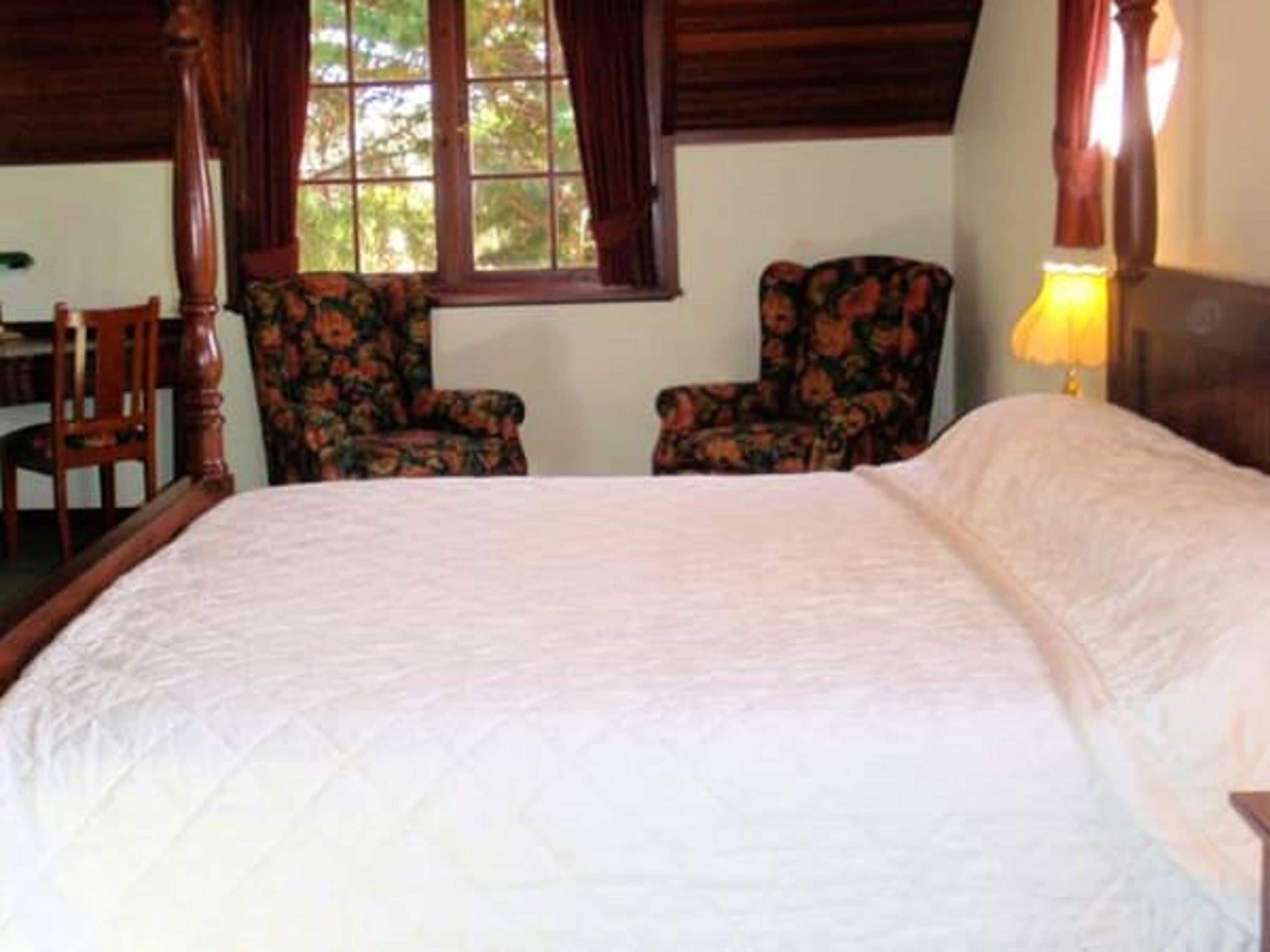 Une chambre à coucher avec un lit à baldaquin, deux fauteuils à motifs floraux, un bureau en bois et une fenêtre avec des rideaux.
