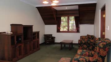 Una habitación con techo inclinado, un armario de madera, un sillón de cuero, un sofá floral y una ventana con cortinas.