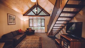 Una sala de estar acogedora con techo de madera, un ventanal grande, una escalera, un sofá, un televisor y una alfombra.