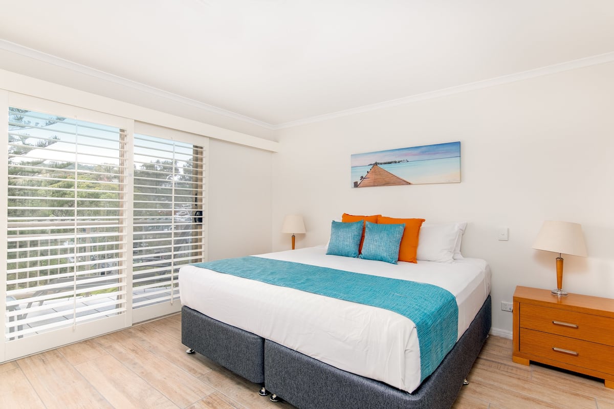 A bedroom with a bed, a nightstand, a window with blinds, and a painting on the wall.