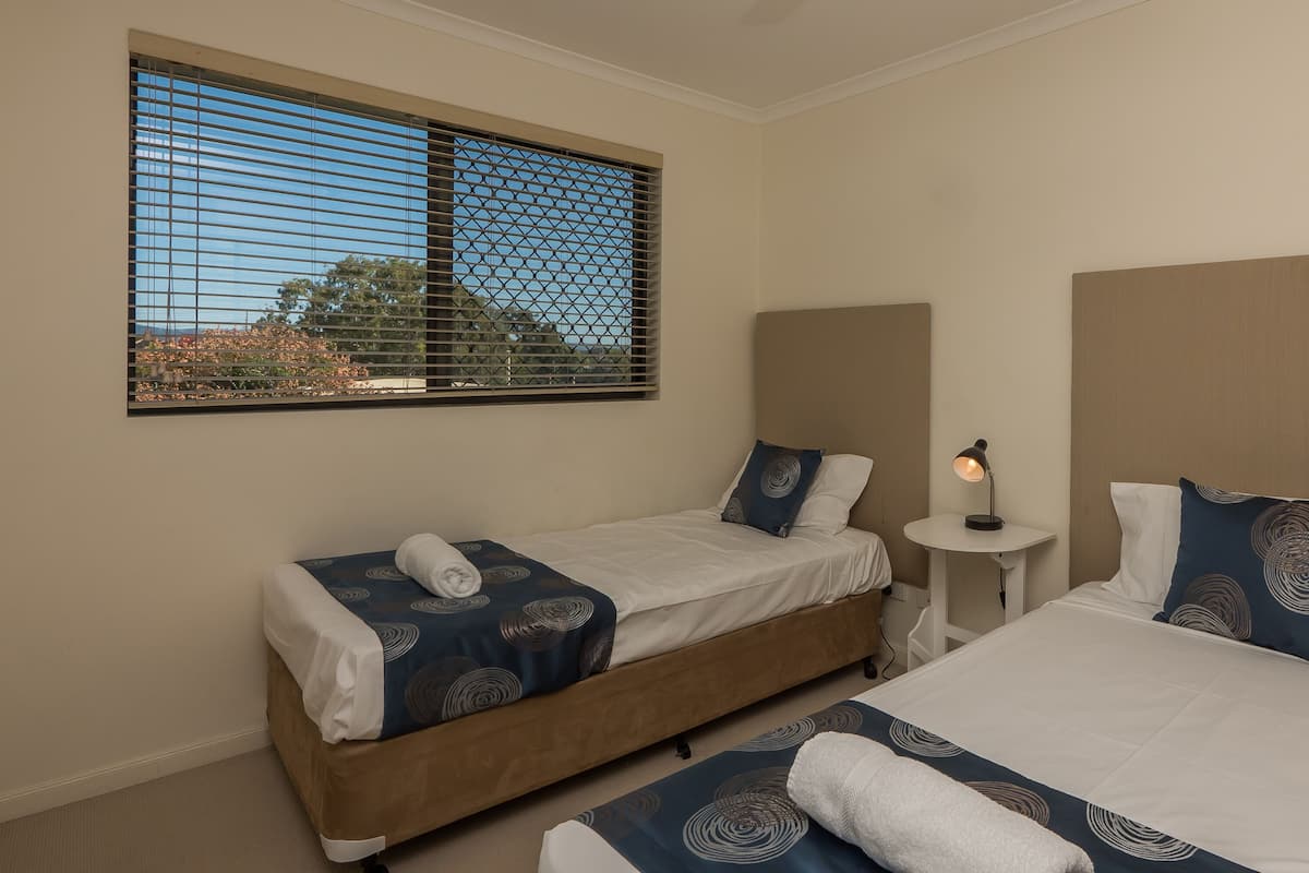 A hotel room with two single beds, a small table with a lamp, and a window with blinds.