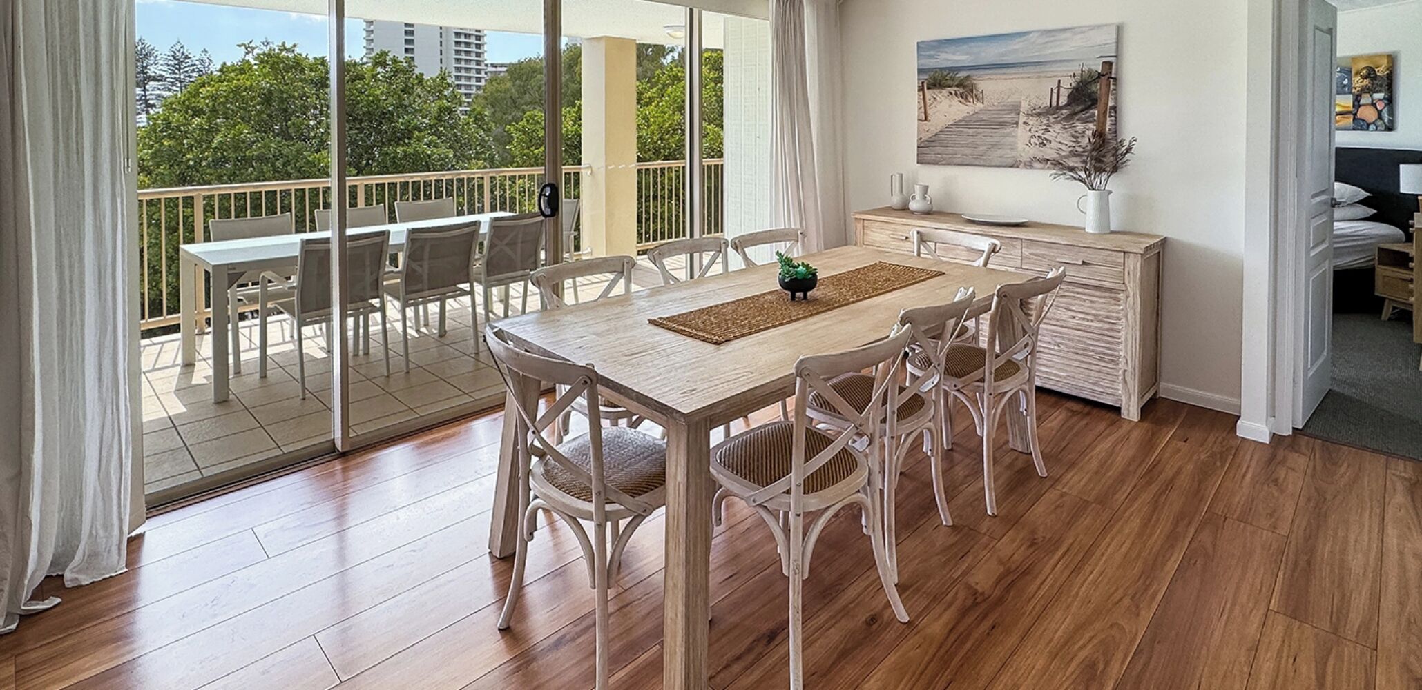 Un comedor con una mesa y sillas de madera, vistas al exterior a través de amplios ventanales y un cuadro en la pared.