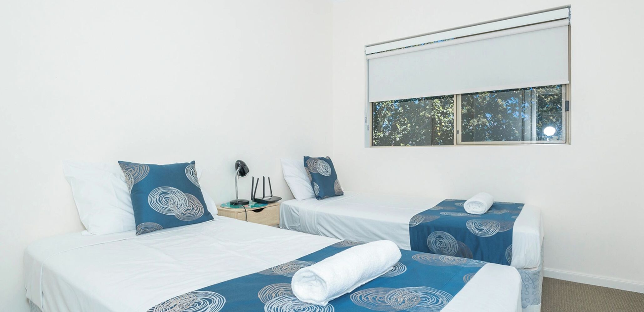 Ein Hotelzimmer mit zwei Betten, jeweils mit blauen, gemusterten Bettdecken und weißen Kissen. Es gibt ein Fenster mit Blick auf Bäume sowie einen Nachttisch mit Lampe und Steckdose.