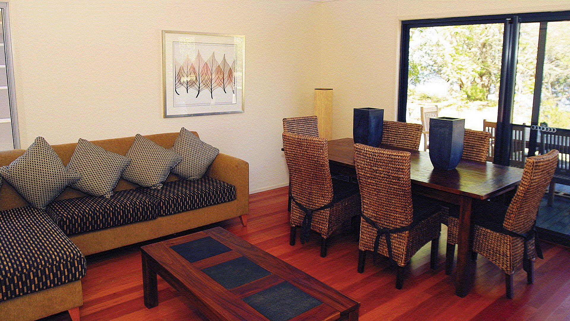 A living room with a sofa, patterned cushions, a wooden coffee table, wicker chairs, a dining table, and a framed picture on the wall.