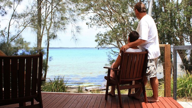 A wooden deck with two chairs and a person standing beside a child. - NRMA Myall Shores Holiday Park (Bombah Point)