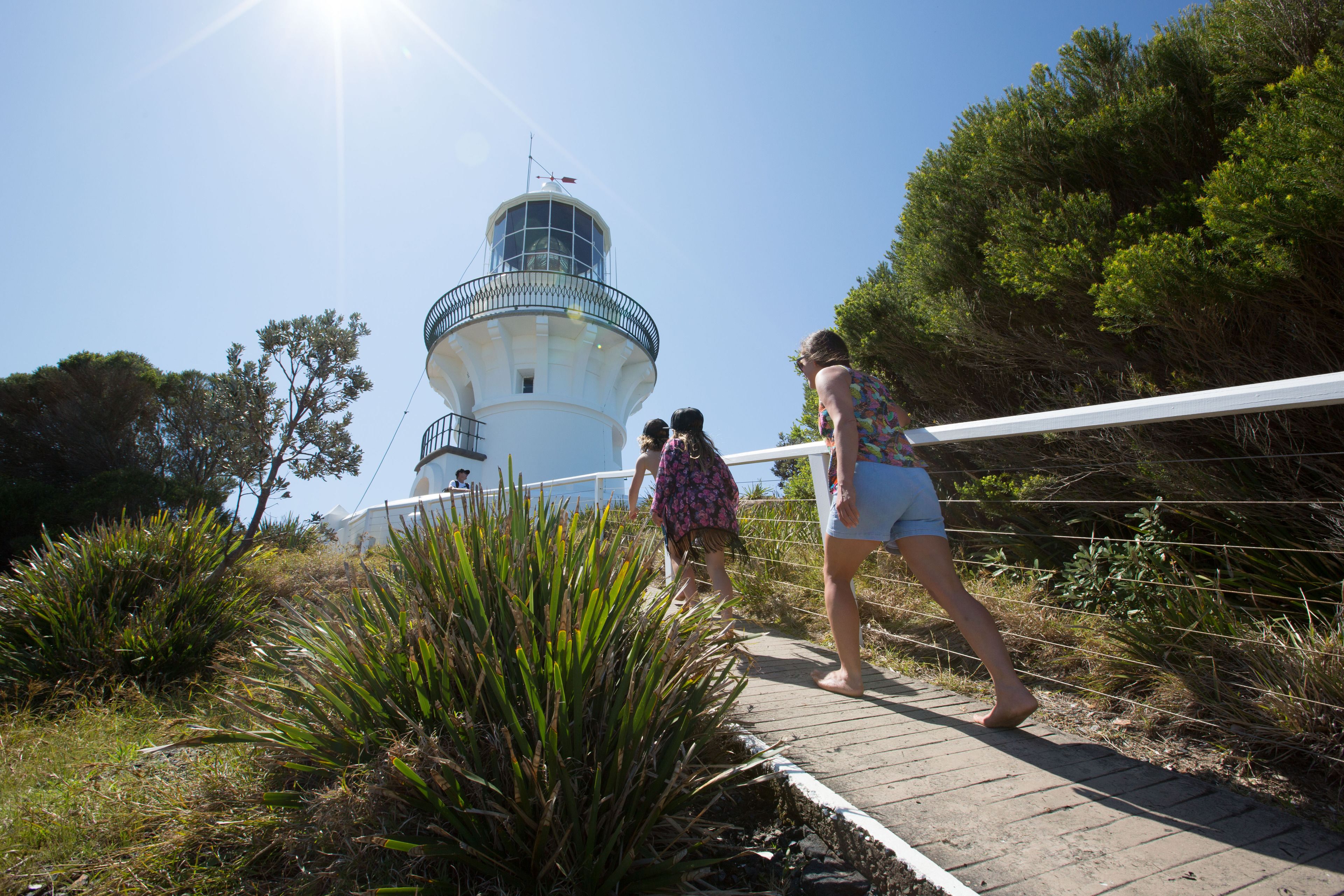A lighthouse with a white exterior and a blue railing, surrounded by greenery and a clear sky.