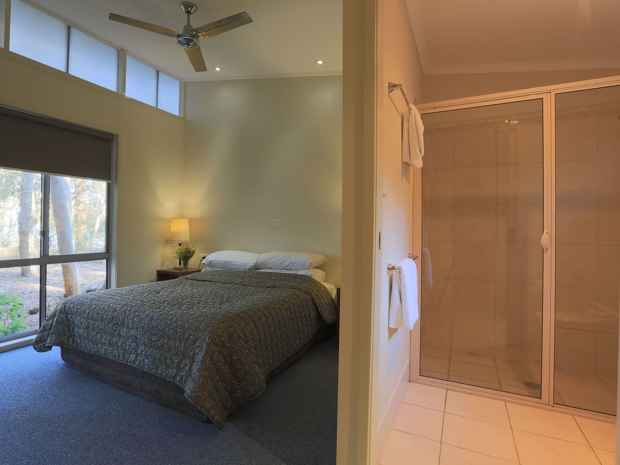 A bedroom with a bed, a nightstand, a lamp, a window with a view of trees, and a shower area.