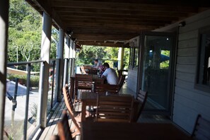 Un espace extérieur aménagé, abrité et doté de tables et de chaises en bois, de grandes fenêtres et d’une vue sur la verdure.