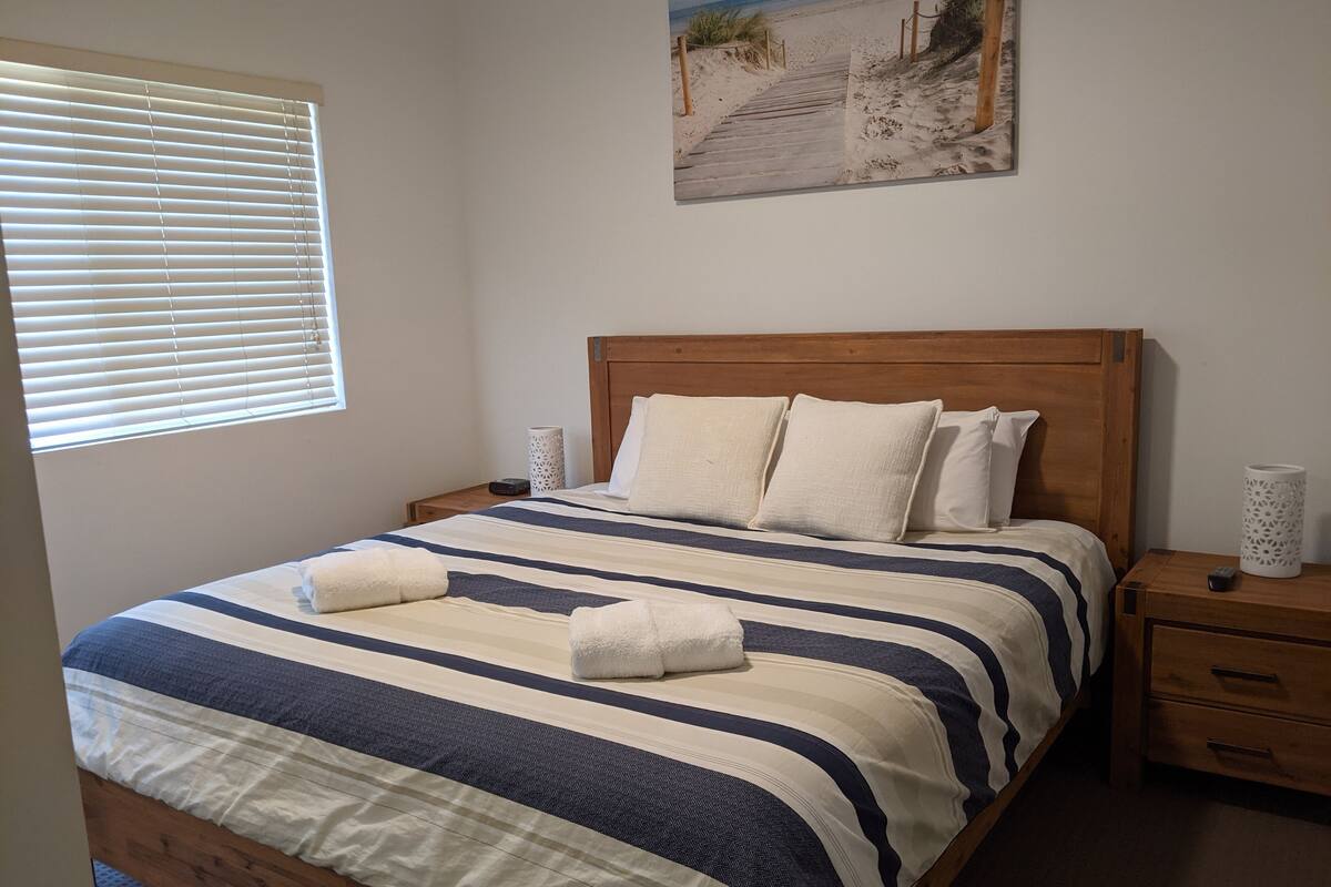 Un dormitorio con cama de madera, ropa de cama a rayas, un cuadro con motivos playeros y una ventana con persianas.