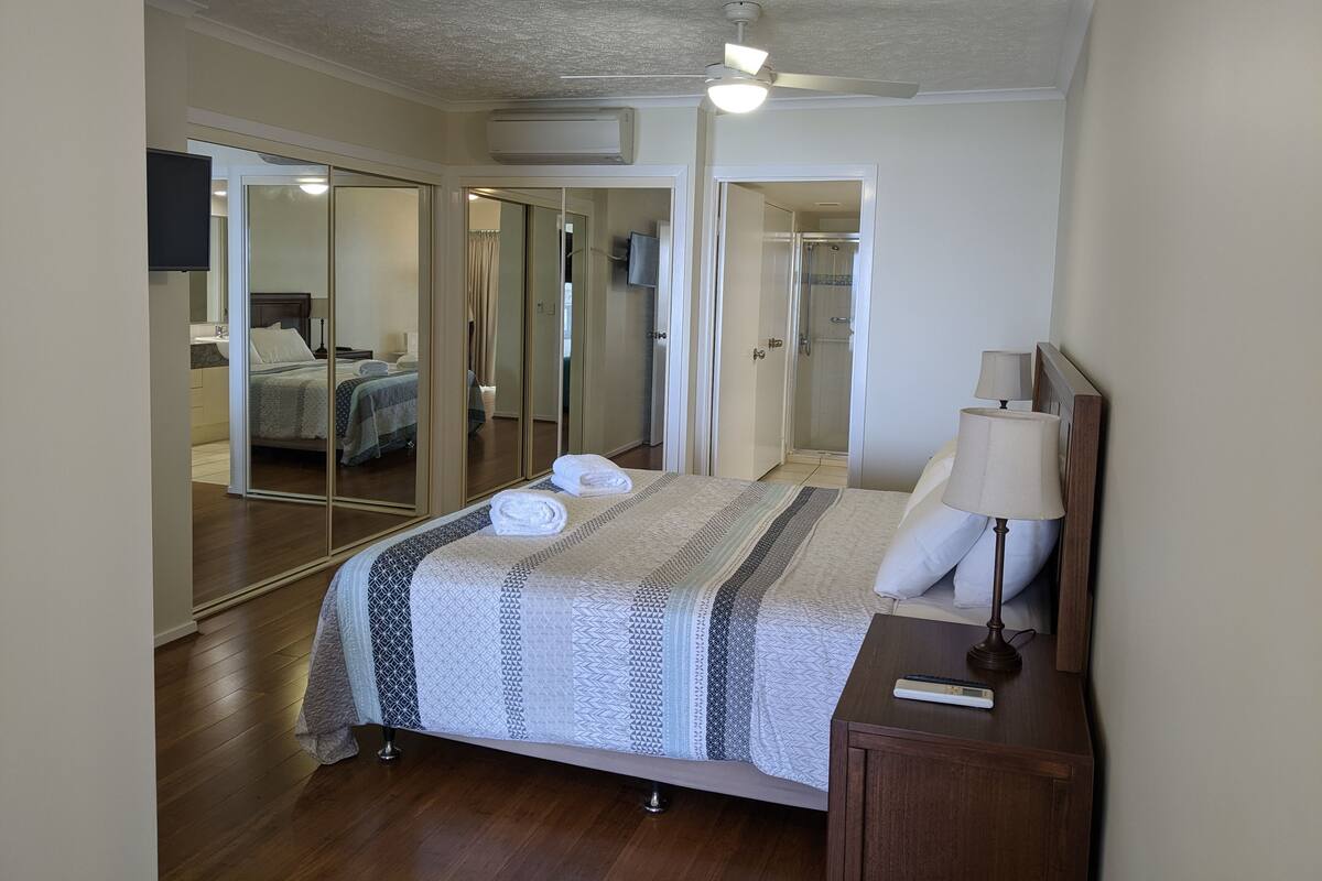 Un dormitorio con cama, mesita de noche con lámpara, espejo, armario y zona de ducha.