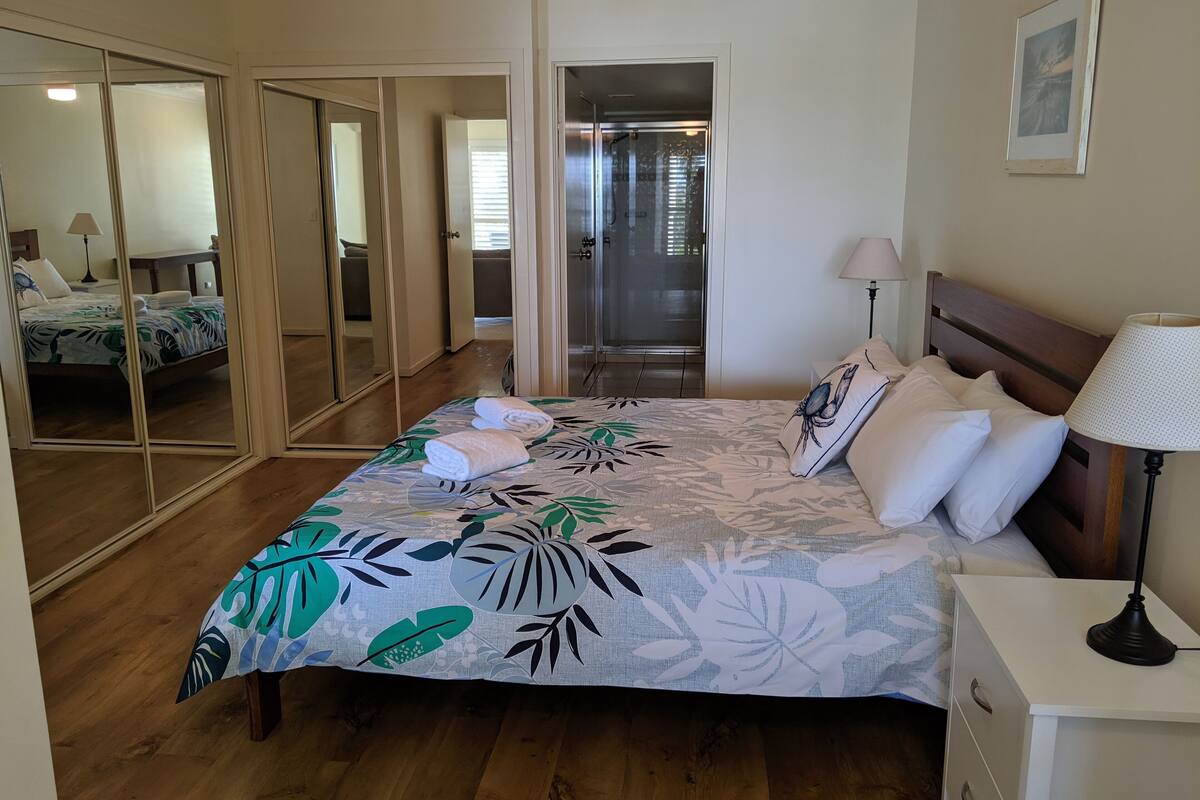 Un dormitorio con cama, mesita de noche, lámpara, espejo y puerta que da al baño.