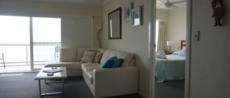 Sala de estar com sofá, mesa de centro e cama com mesa de cabeceira. Há uma janela grande com vista para o oceano.