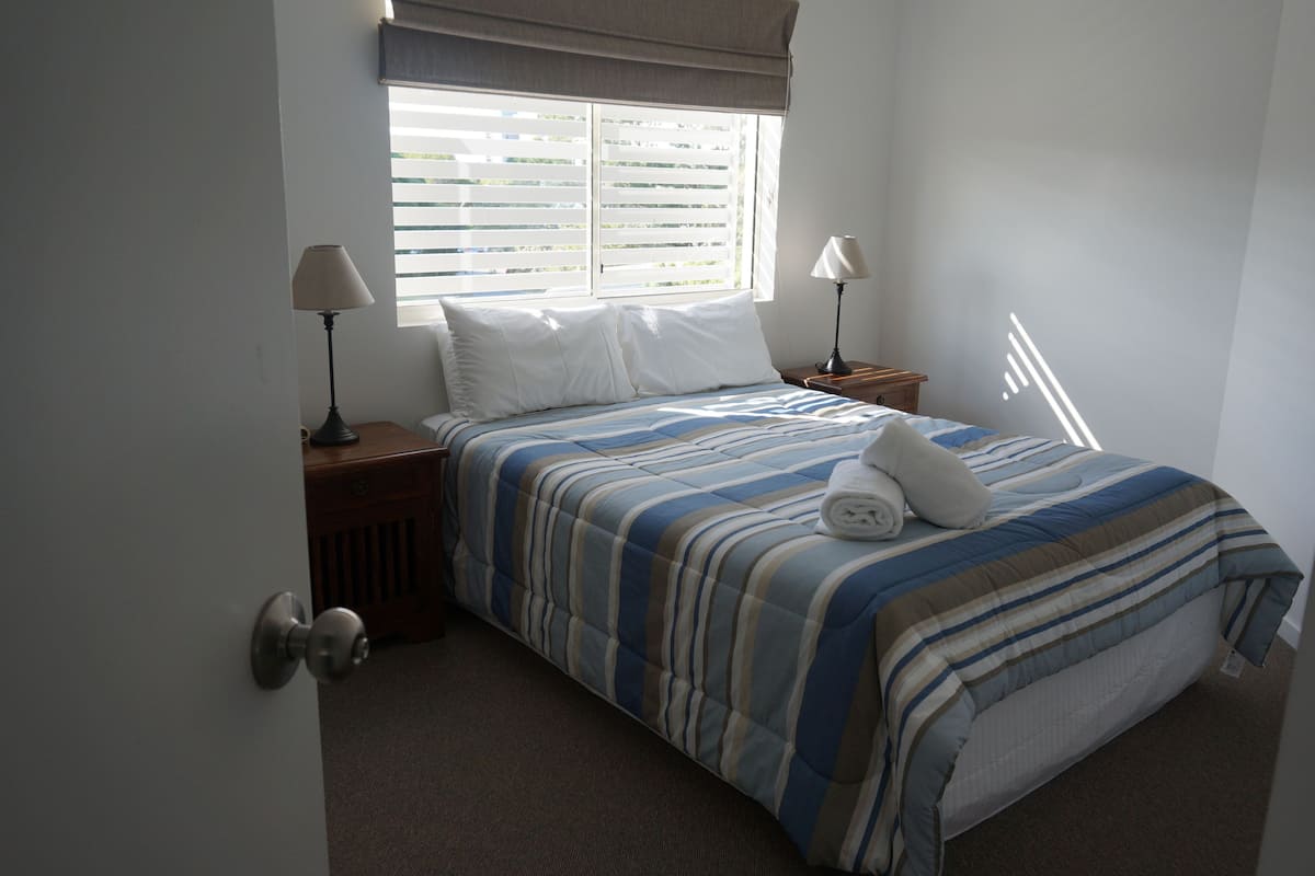 Una cama bien tendida con ropa de cama a rayas, dos lámparas de noche y una ventana con persianas.