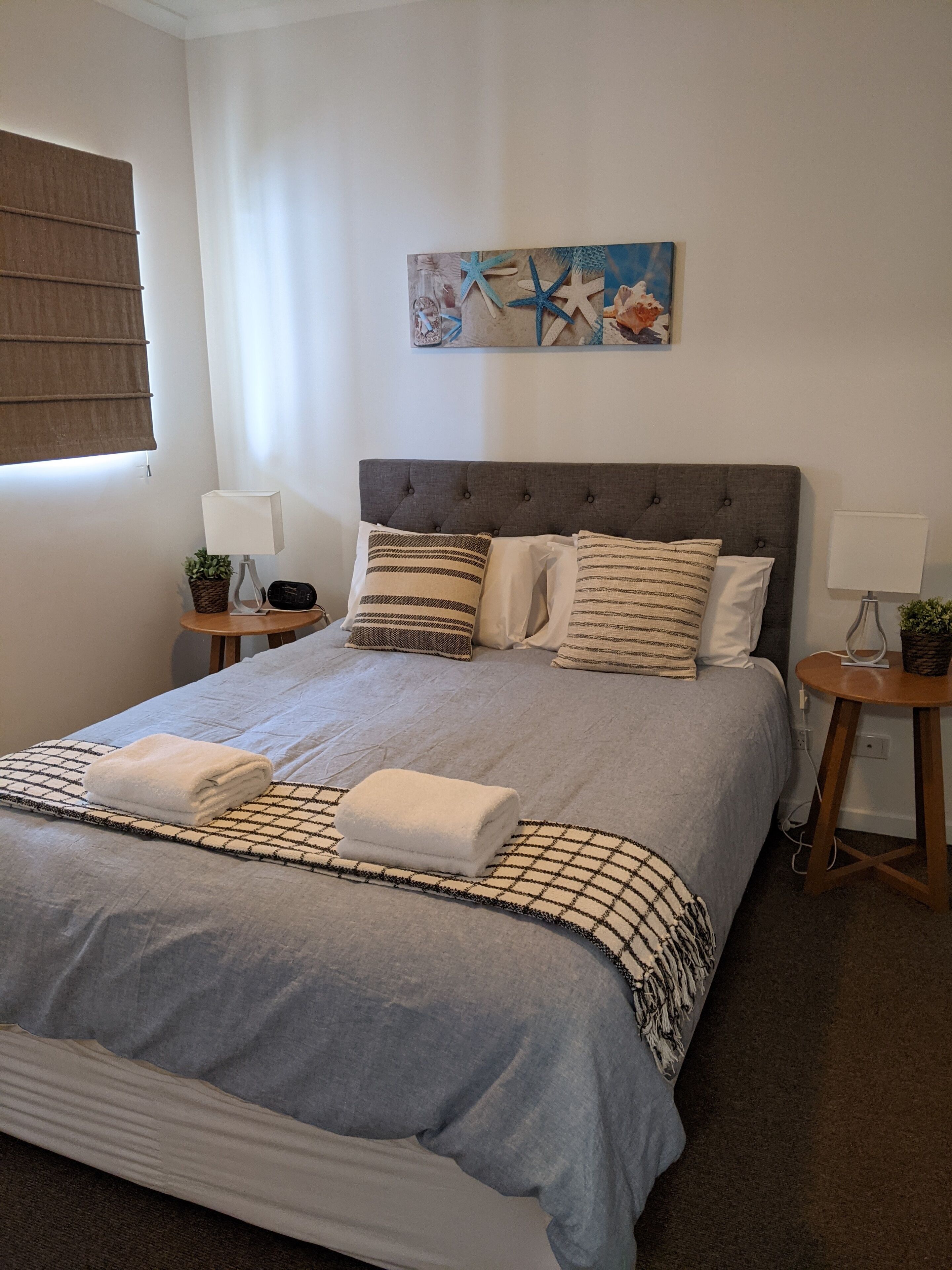 A neatly made bed with a grey duvet, multiple pillows, and two bedside tables with lamps and small plants.